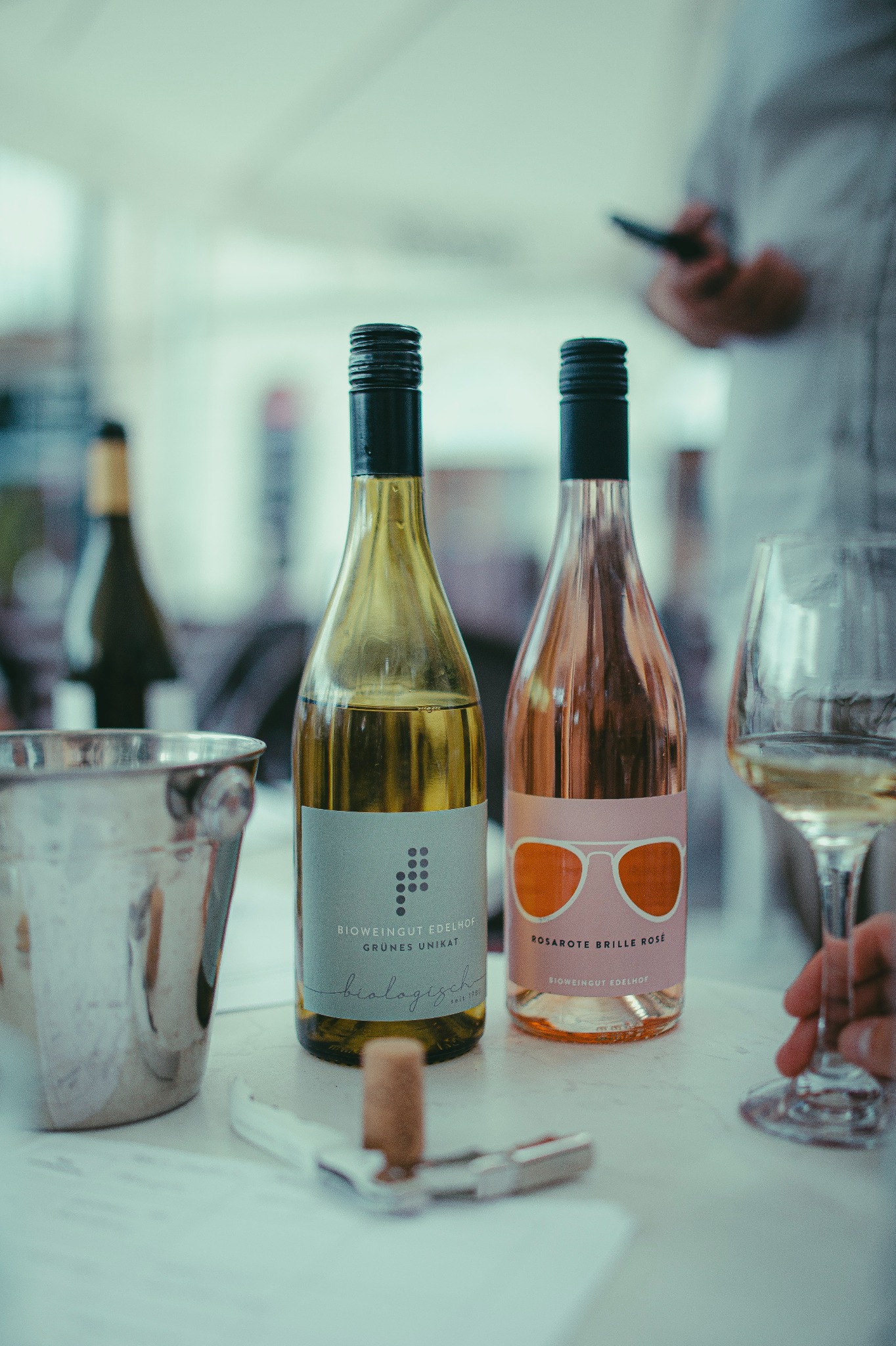 Dwie butelki wina (białe i różowe) na stole z korkociągiem i kieliszkiem. W tle osoba z telefonem. Etykiety z grafiką okularów i kropek.