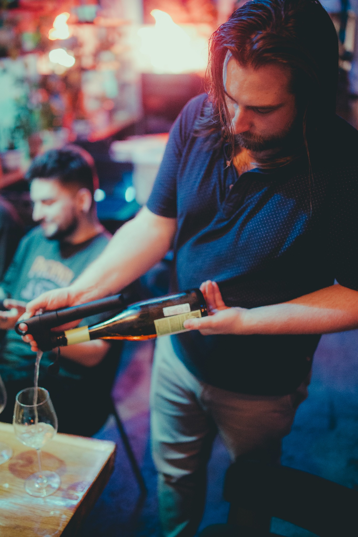 Barman nalewa białe wino z butelki do kieliszka na drewnianym blacie, w tle siedzący mężczyzna i dekoracje świetlne tworzą kameralny nastrój.