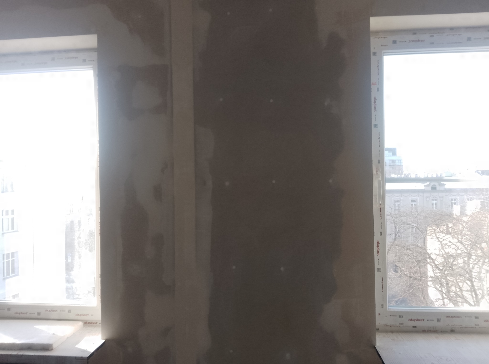 Ściana po nałożeniu gładzi szpachlowej, widoczne dwa okna z białymi ramami. Przez okna widać budynki i drzewa. Ściana przygotowana do dalszych prac.