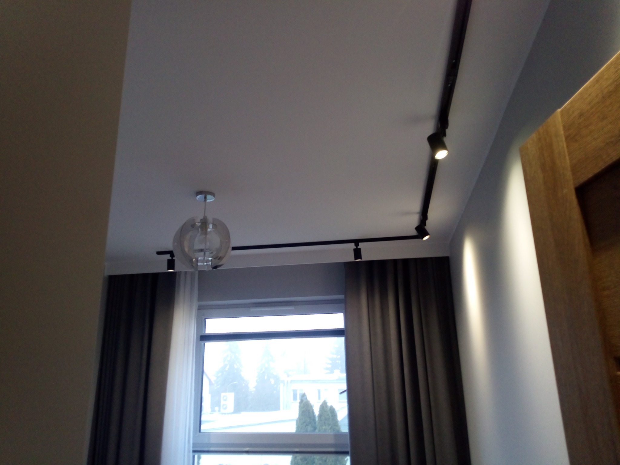 Nowoczesne oświetlenie sufitowe z reflektorami na szynie i designerską lampą wiszącą, widok na okno z zasłonami w tle. Minimalistyczny design wnętrza.