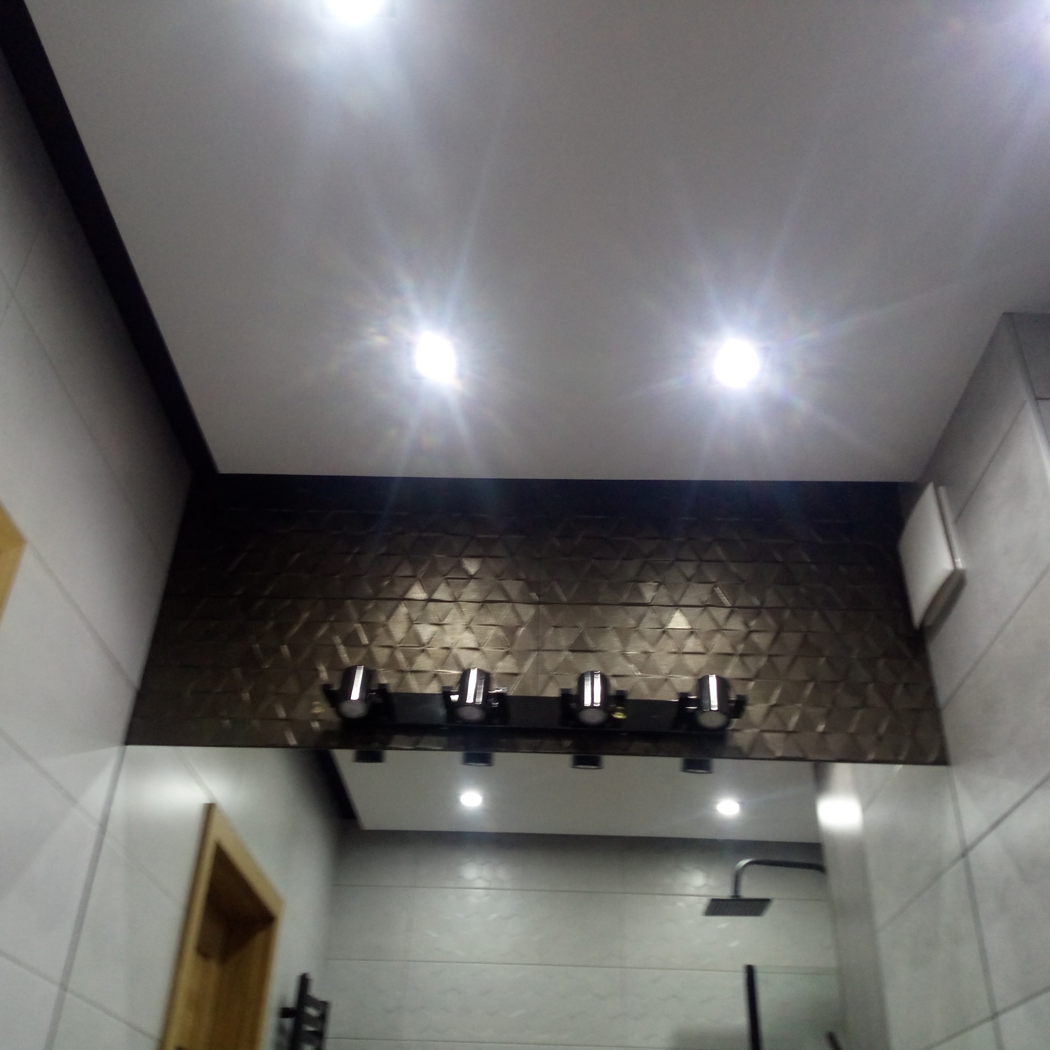 Wykończona łazienka z oświetleniem punktowym, brązową mozaiką i szarymi płytkami. Widoczny fragment kabiny prysznicowej i sufit podwieszany.