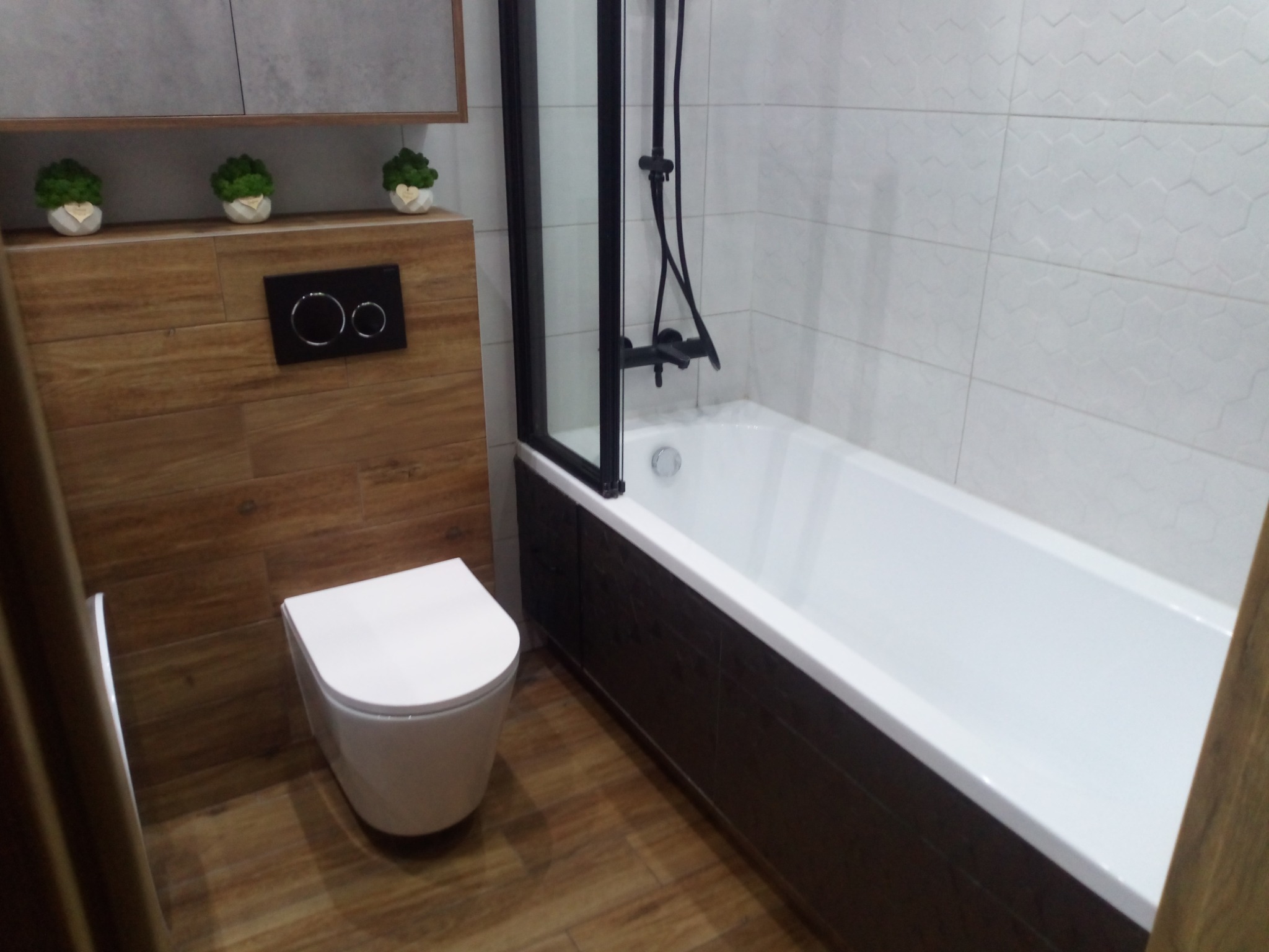 Nowoczesna łazienka po remoncie: biała wanna z czarną armaturą i parawanem, podwieszana toaleta, drewnopodobne płytki i dekoracyjne rośliny w doniczkach.