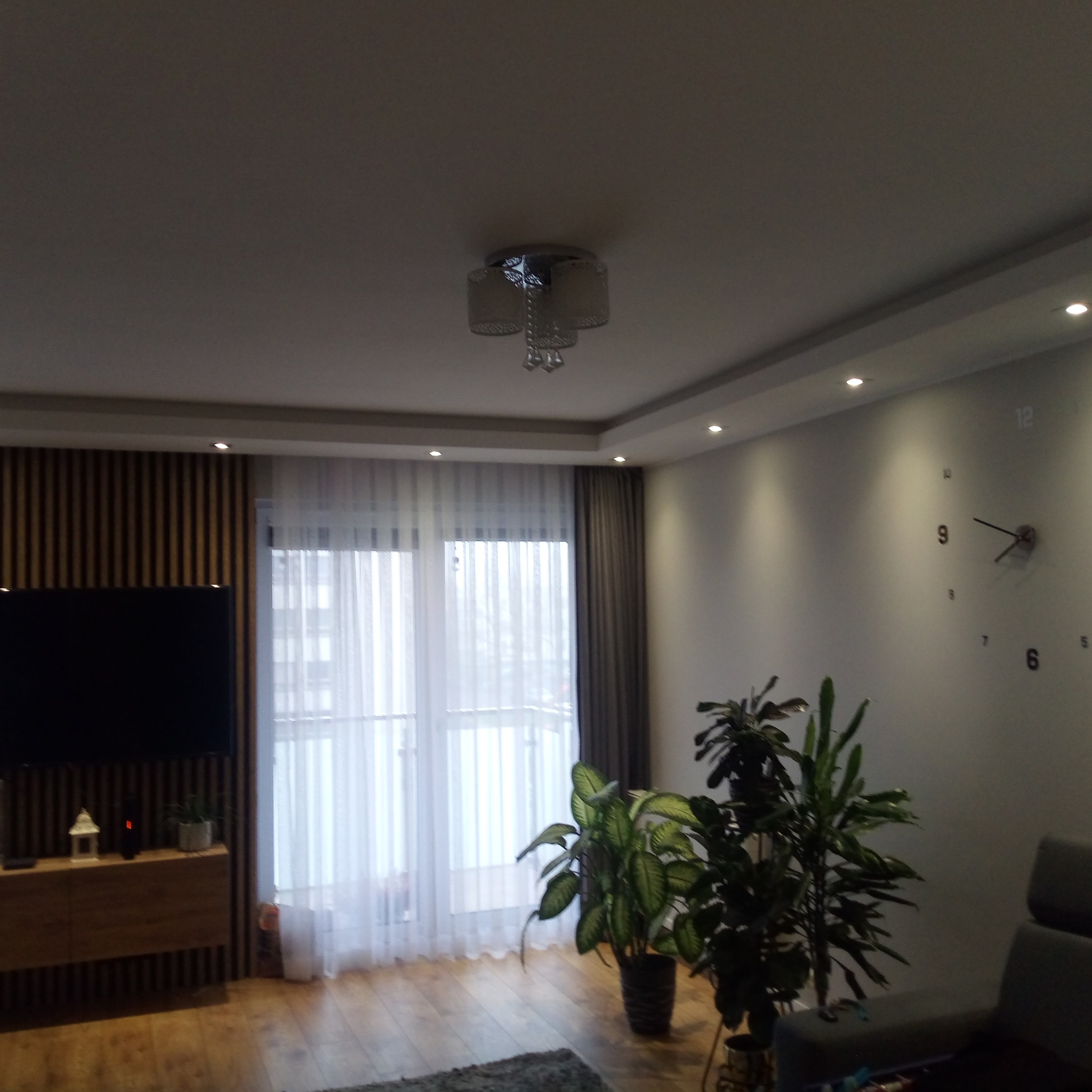 Nowoczesny salon z drewnianą ścianą TV, jasnymi zasłonami, zegarem ściennym i roślinami doniczkowymi. Oświetlenie sufitowe punktowe i lampa wisząca.