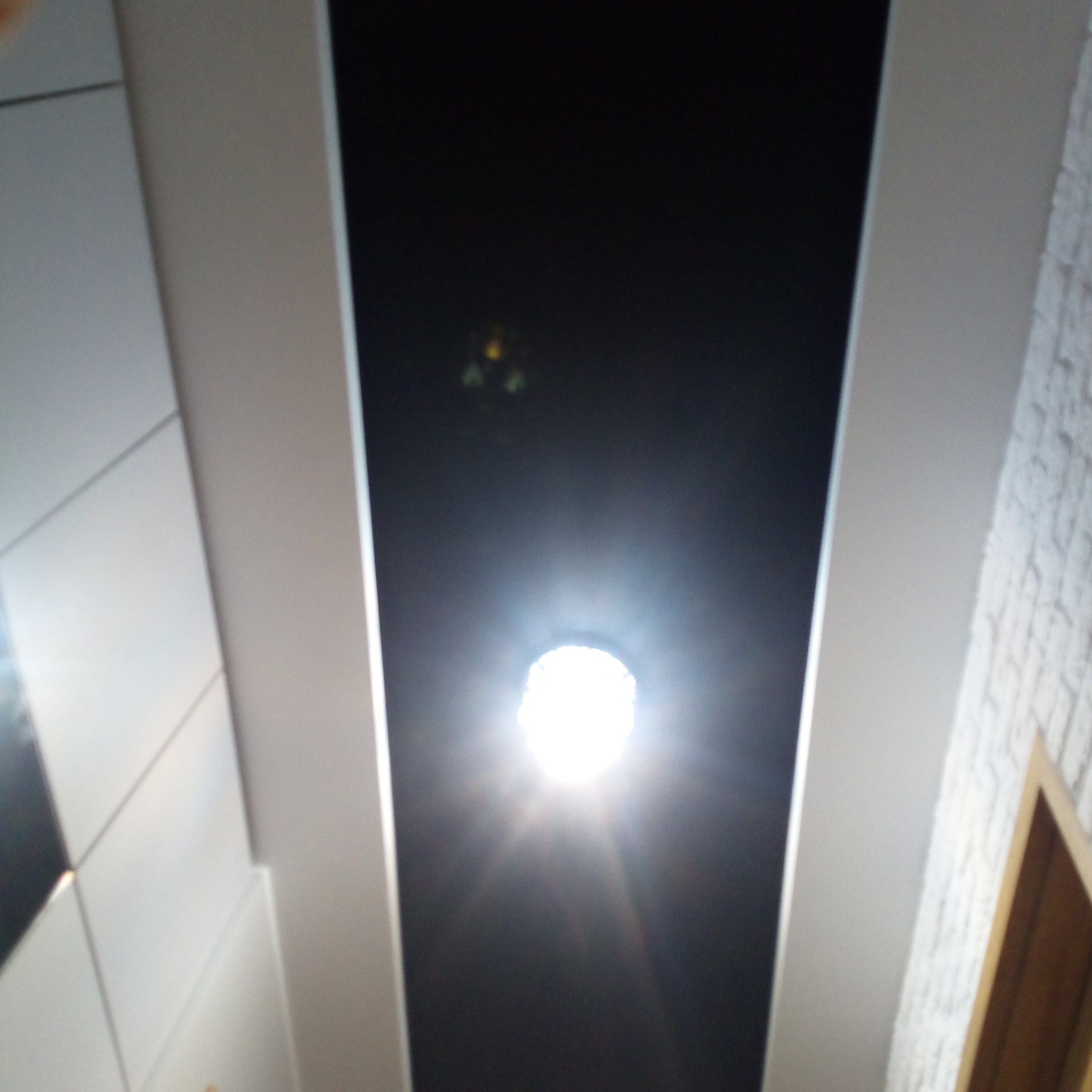 Nowoczesny sufit podwieszany z czarnym panelem i punktowym oświetleniem LED, widziany z dołu. Białe panele boczne i fragment ściany z fakturą.