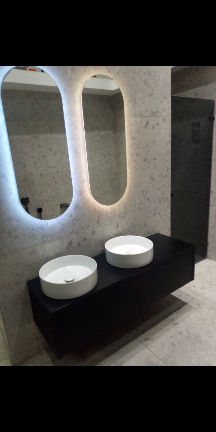 Nowoczesna łazienka z dwoma okrągłymi umywalkami nablatowymi na czarnej szafce, podświetlane lustra o zaokrąglonym kształcie i szklana kabina prysznicowa.