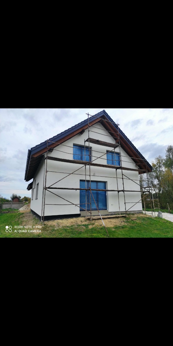 Dwukondygnacyjny dom z rusztowaniem, okna zabezpieczone folią, świeżo pomalowana elewacja na biało, widok z zewnątrz, pochmurne niebo w tle.