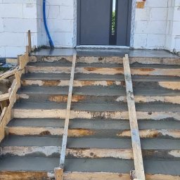 Paweł Bakowski - Betonowe schody wejściowe w trakcie budowy, z drewnianym szalunkiem i świeżym betonem. Widoczne narzędzia i fragment elewacji budynku z białego bloczka.