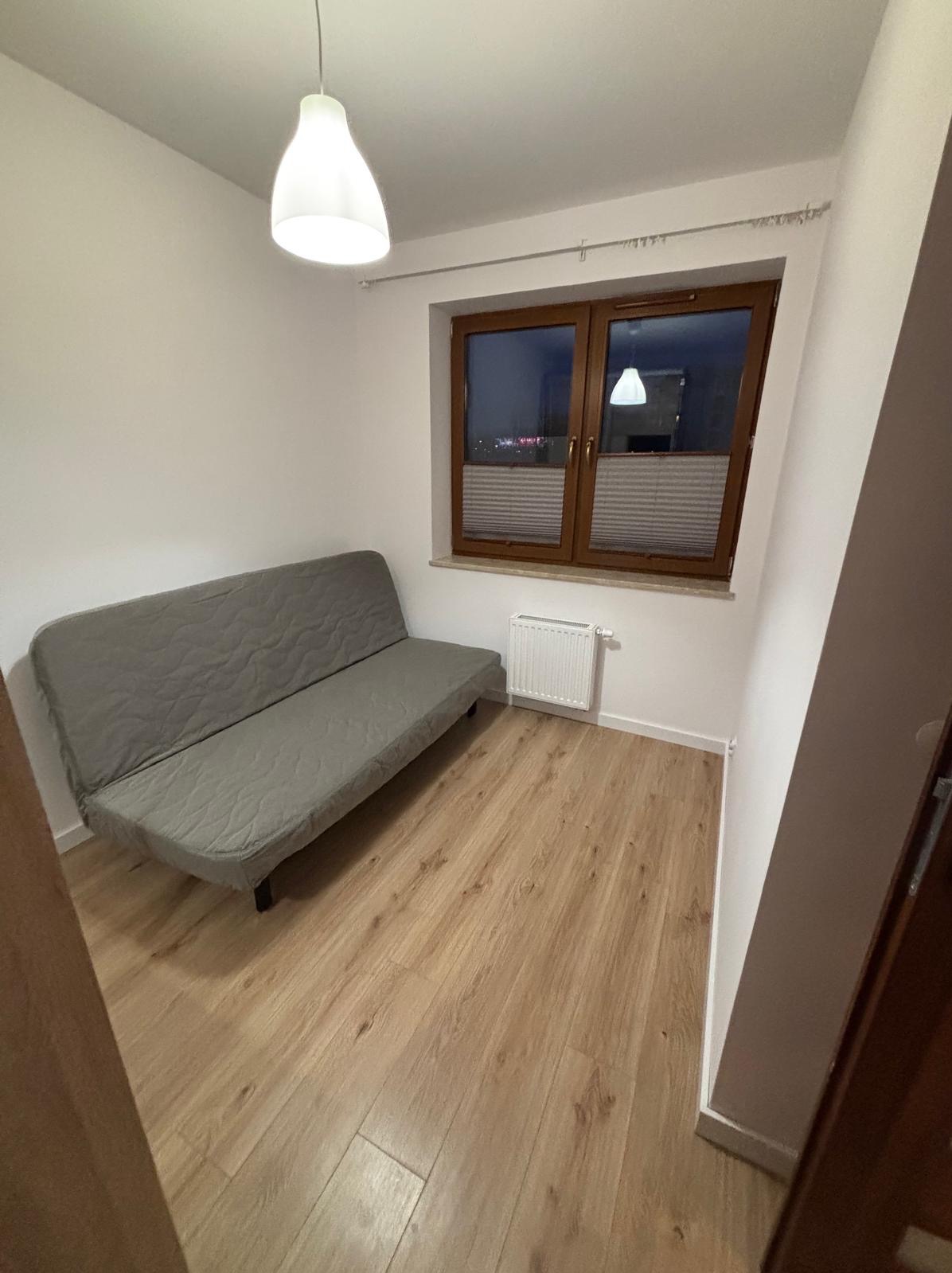 Pokój z szarą sofą pod oknem z żaluzjami, drewniana podłoga, białe ściany i sufit, lampa wisząca. Widok na miasto za oknem.