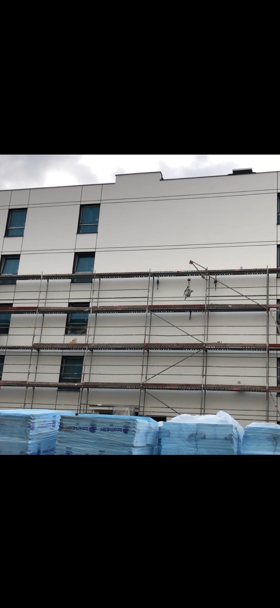 Budynek w trakcie ocieplania, widoczne rusztowania i stosy styropianu. Elewacja z panelami, okna z ciemnymi ramami. Ujęcie z dołu, pochmurne niebo.