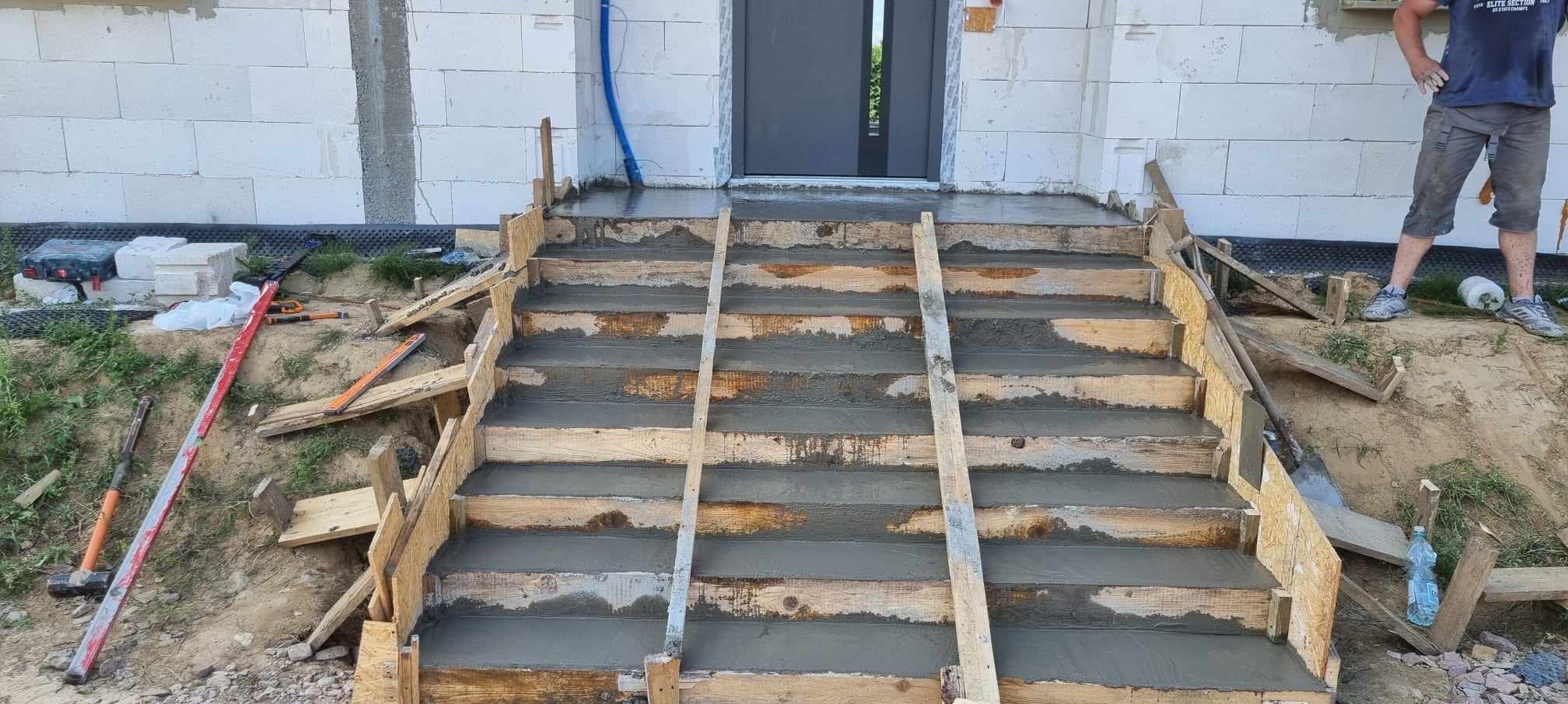 Betonowe schody wejściowe w trakcie budowy, z drewnianym szalunkiem i świeżym betonem. Widoczne narzędzia i fragment elewacji budynku z białego bloczka.