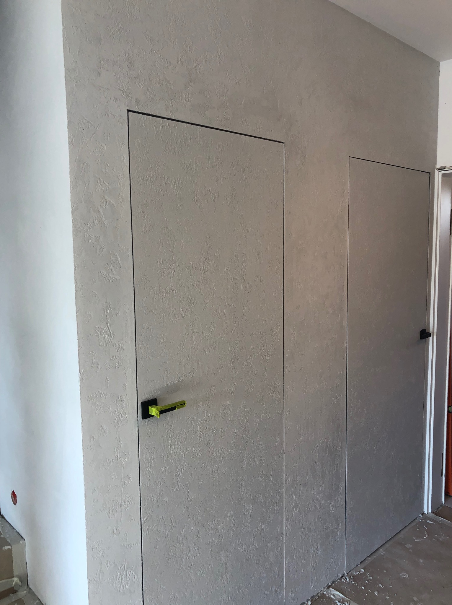 Dwa ukryte wejścia w ścianie z dekoracyjną, szarą strukturą. Minimalistyczne drzwi z czarnymi obramowaniami i zielonymi akcentami na klamkach. Wykończenie wnętrza.