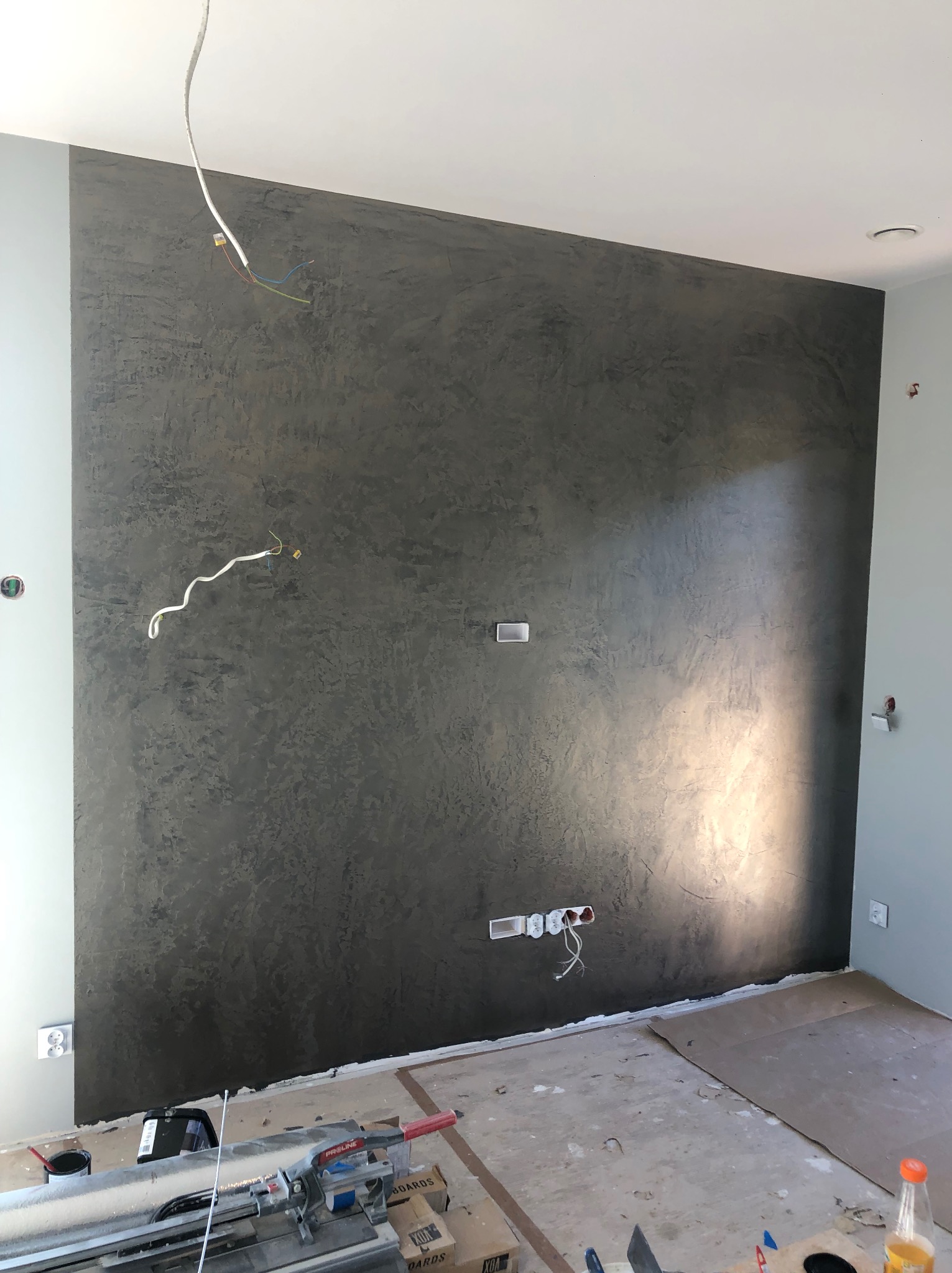 Ściana w pokoju po nałożeniu dekoracyjnej, ciemnej gładzi w stylu weneckim. Widoczne puszki elektryczne i narzędzia na podłodze.