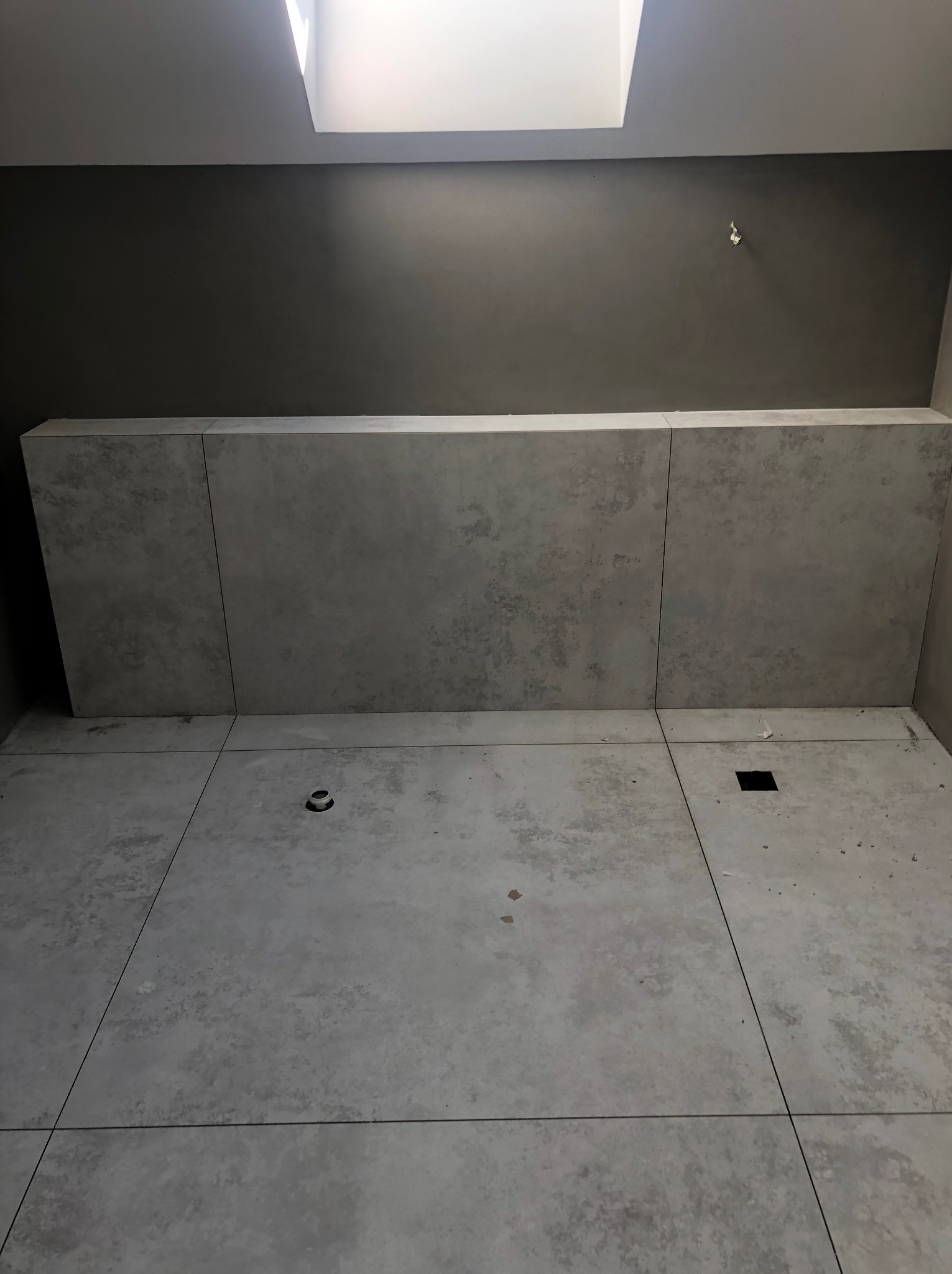 Minimalistyczna łazienka w szarościach: płytki imitujące beton na podłodze i ścianie, sufit z oknem dachowym. Widoczne odpływy liniowe i punktowe.