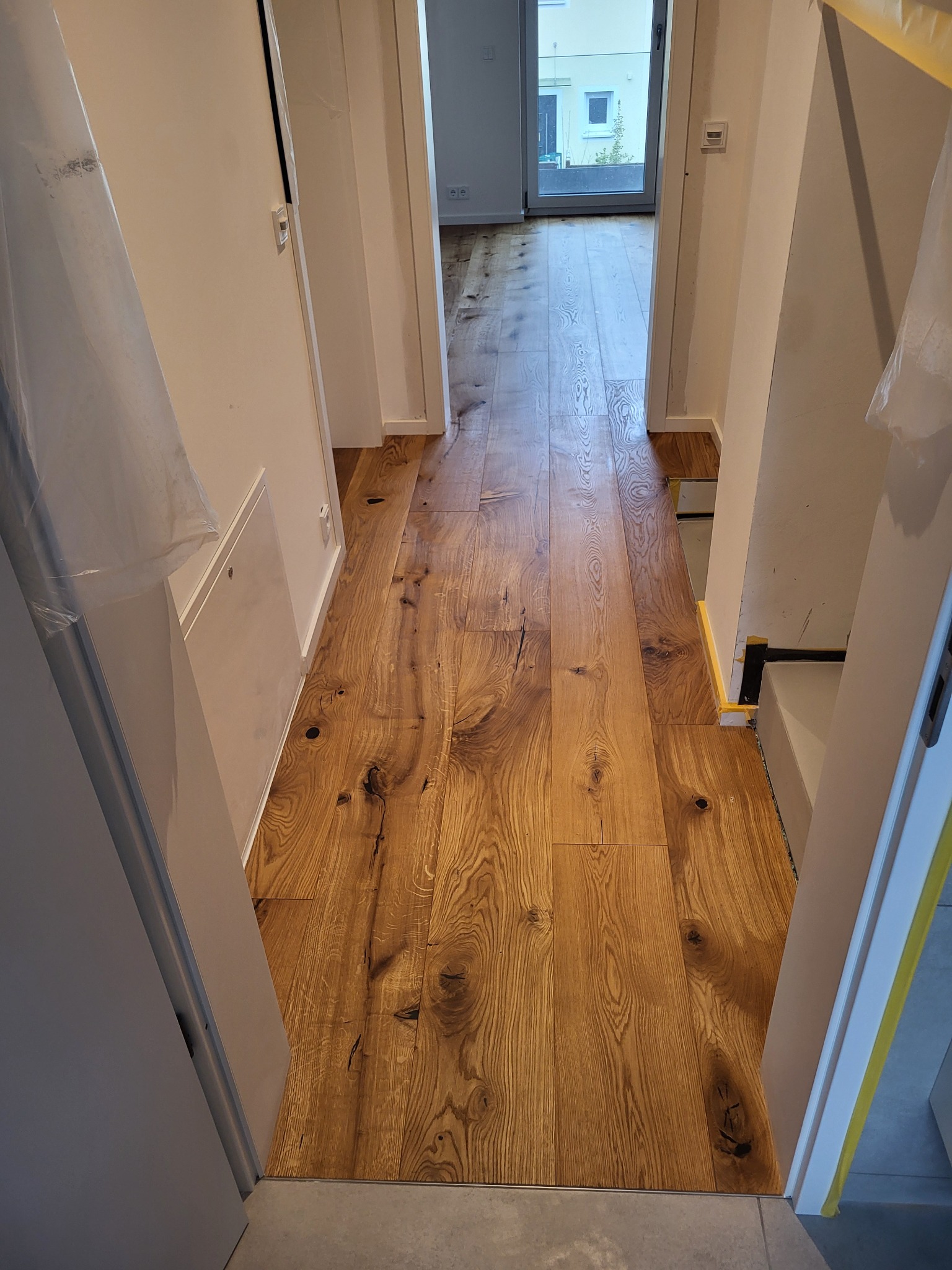 Ułożony parkiet dębowy w korytarzu, widok z góry. Deski z wyraźnym usłojeniem i sękami. Ściany białe, zabezpieczone folią malarską. Widoczne listwy przypodłogowe.
