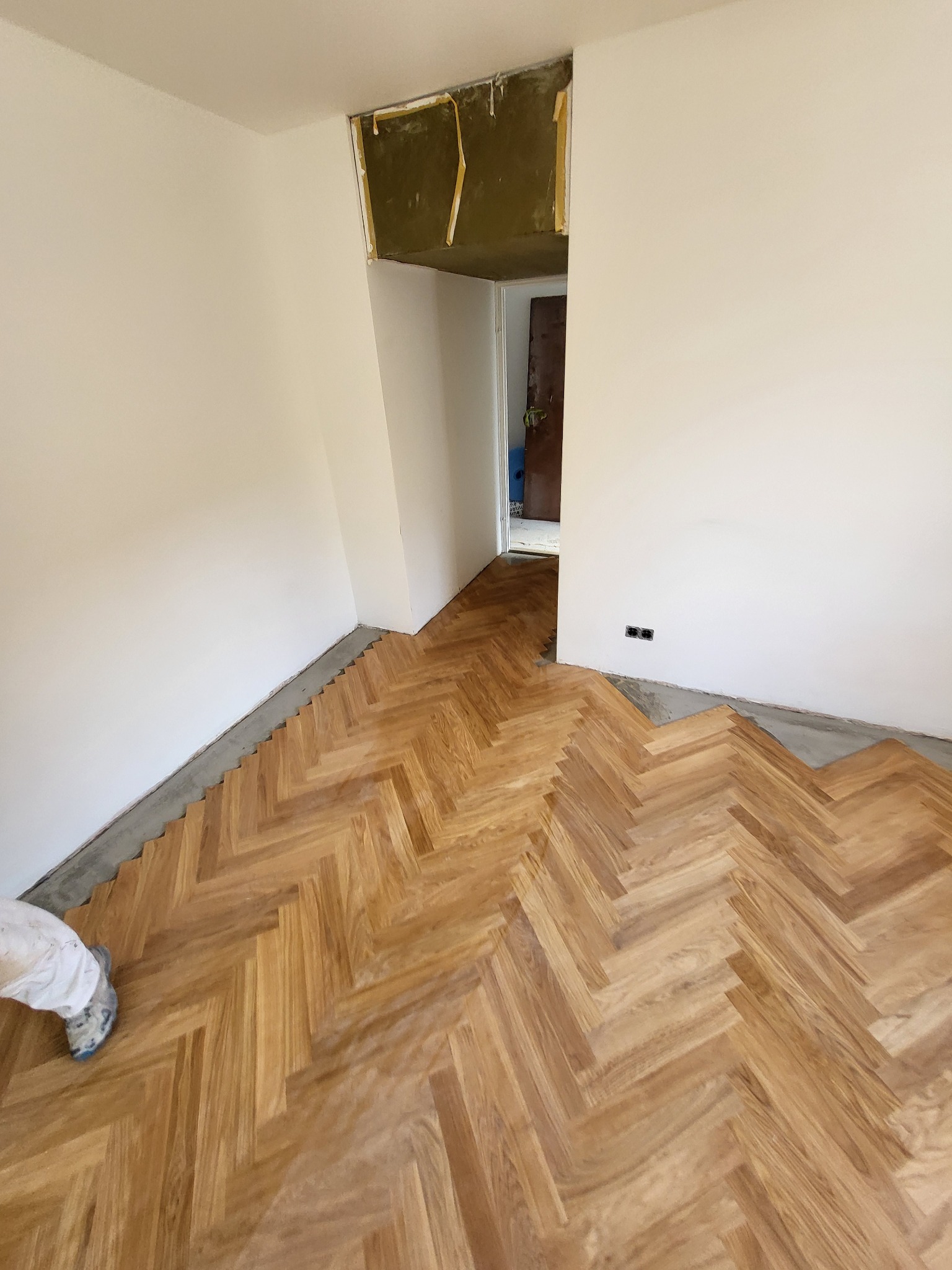 Układanie parkietu w jodełkę w jasnym pokoju. Widoczne fragmenty białych ścian i przygotowane podłoże. W tle widoczne wejście do innego pomieszczenia.
