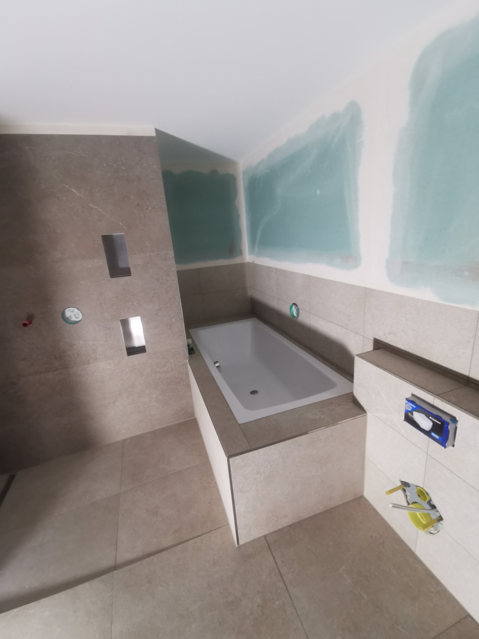 Remont łazienki w trakcie: obudowana wanna, płytki w odcieniach beżu, przygotowane ściany do malowania, widoczne instalacje. Minimalistyczny design.