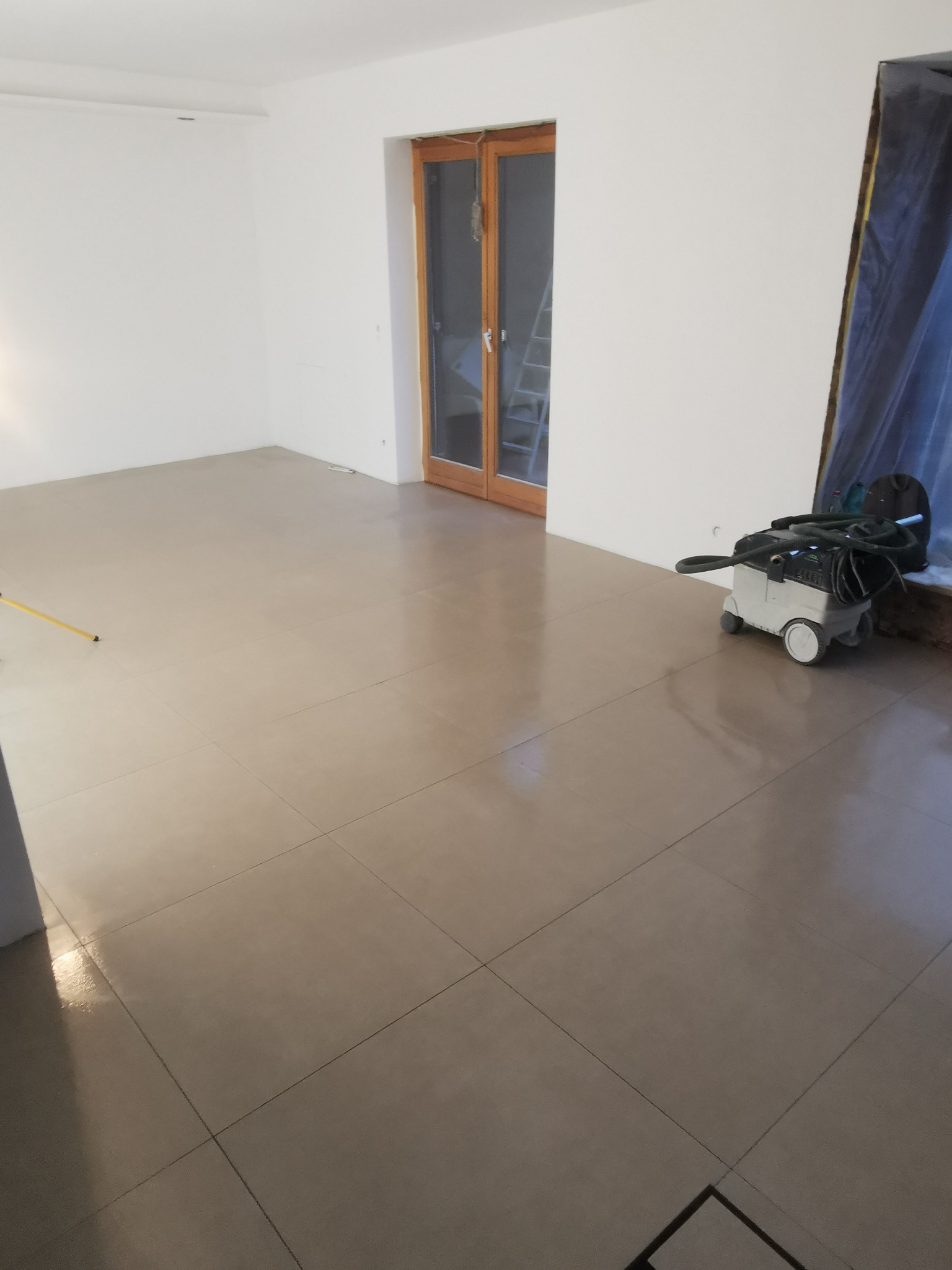 Błyszcząca, świeżo ułożona podłoga z płytek w pustym pokoju z białymi ścianami. Widoczny odkurzacz przemysłowy i drewniane drzwi z drabinką w tle.