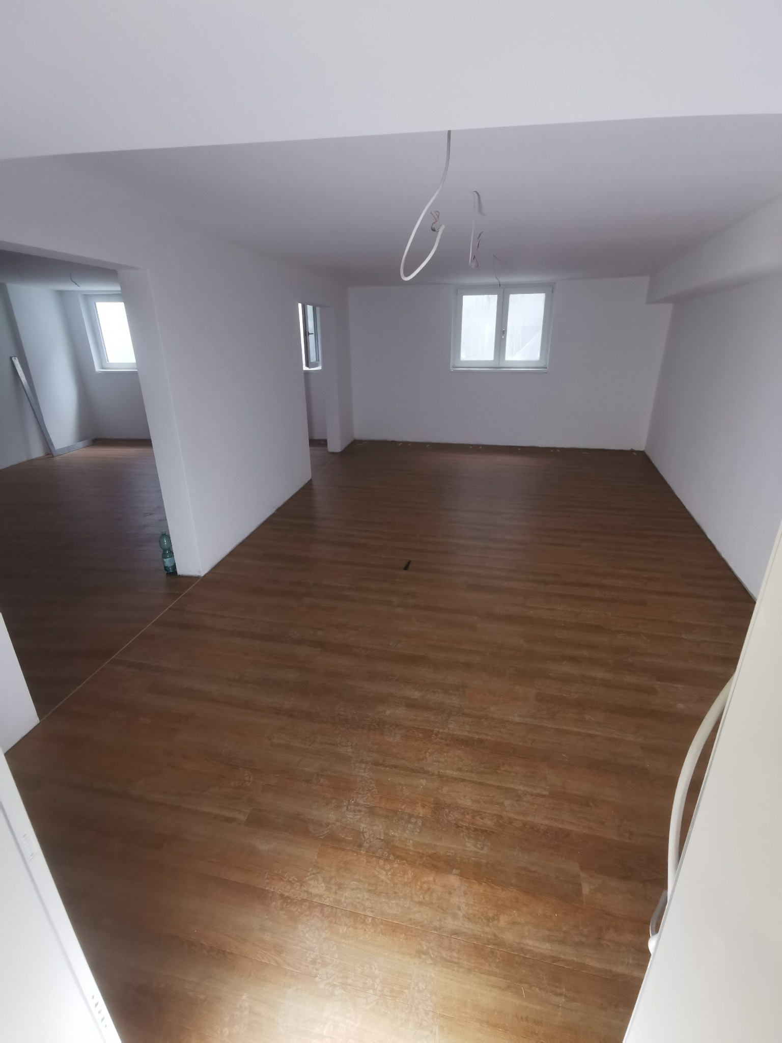 Jasne, przestronne wnętrze po remoncie: świeżo pomalowane ściany, brązowa podłoga imitująca drewno, okna wpuszczające naturalne światło. Widoczne kable zwisające z sufitu.