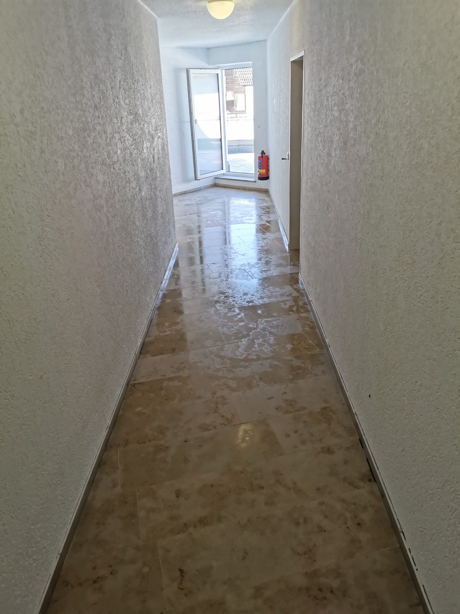 Korytarz po remoncie: beżowe płytki podłogowe z delikatnym wzorem, ściany o fakturze baranka w jasnym odcieniu, widoczne drzwi i okno w oddali.