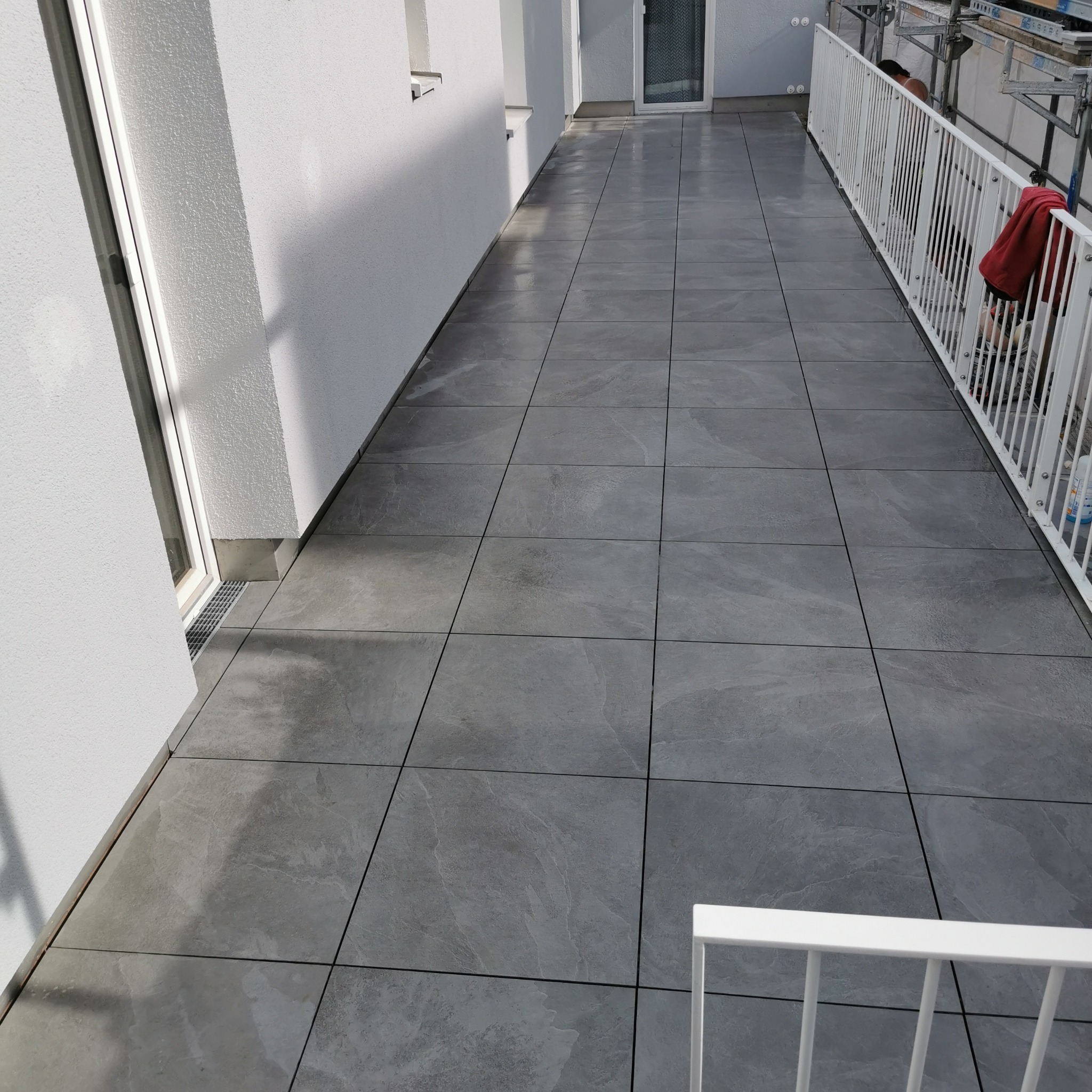 Wyłożony szarymi płytkami taras z białą balustradą. Widoczne detale: fuga, faktura płytek imitująca kamień. Częściowo widoczne rusztowanie w tle.