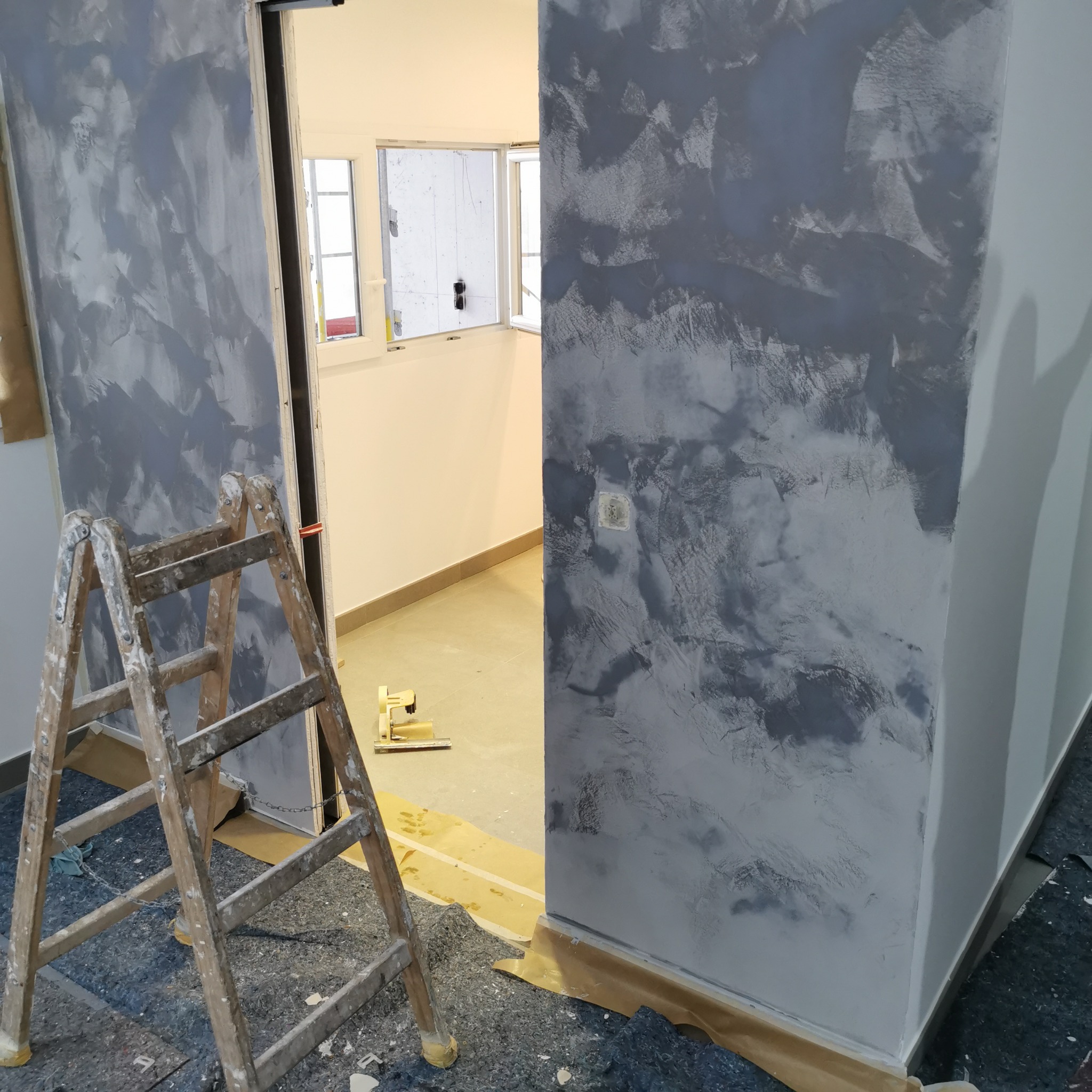 Ściana z efektem dekoracyjnym w odcieniach szarości, drabina malarska i zabezpieczona podłoga. Widok na jasne pomieszczenie z oknem. Prace wykończeniowe.