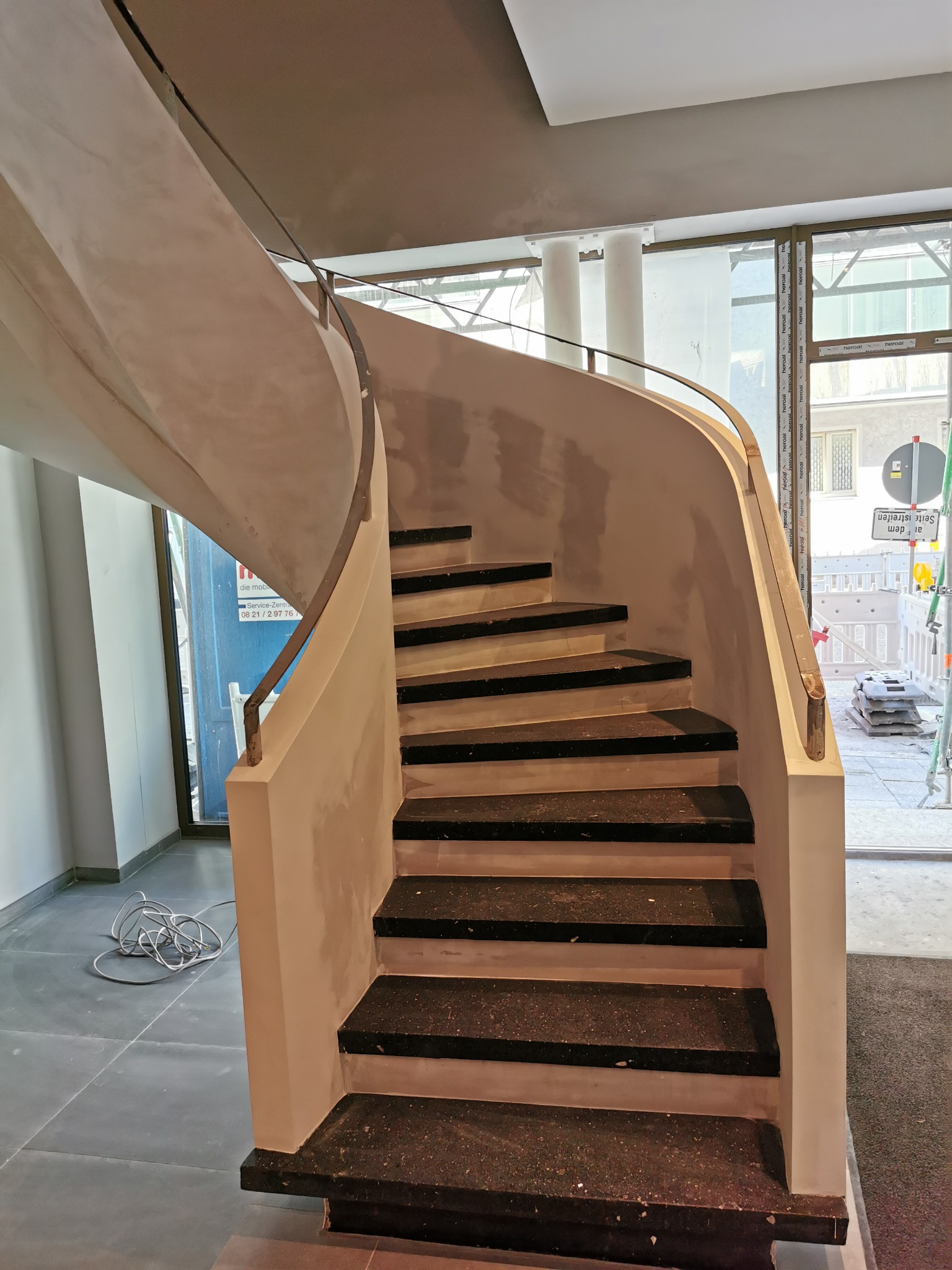 Spiralne schody z czarnymi stopniami i jasnymi ścianami, wykończone metalową poręczą. Widoczne ślady szpachlowania na ścianach.