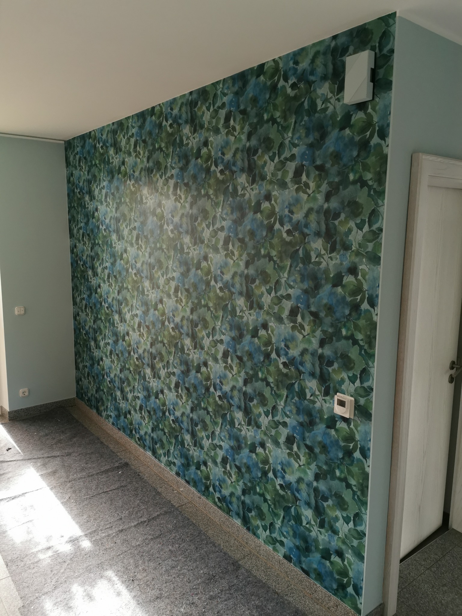 Ściana z wzorem liści w odcieniach zieleni i błękitu, świeżo po malowaniu, z widocznym fragmentem drzwi i elementami instalacji elektrycznej. Jasne, naturalne oświetlenie.