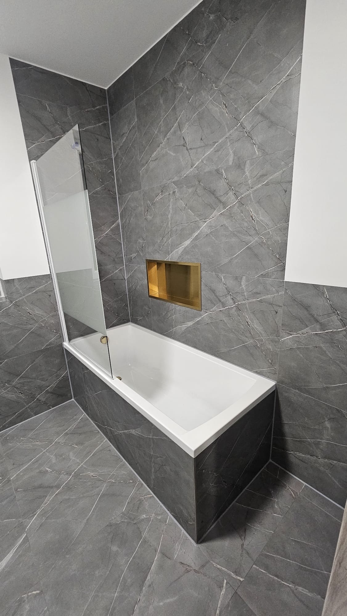 Wykończona łazienka z wanną obudowaną szarymi płytkami imitującymi marmur, szklanym parawanem i złotą wnęką w ścianie. Nowoczesny design.