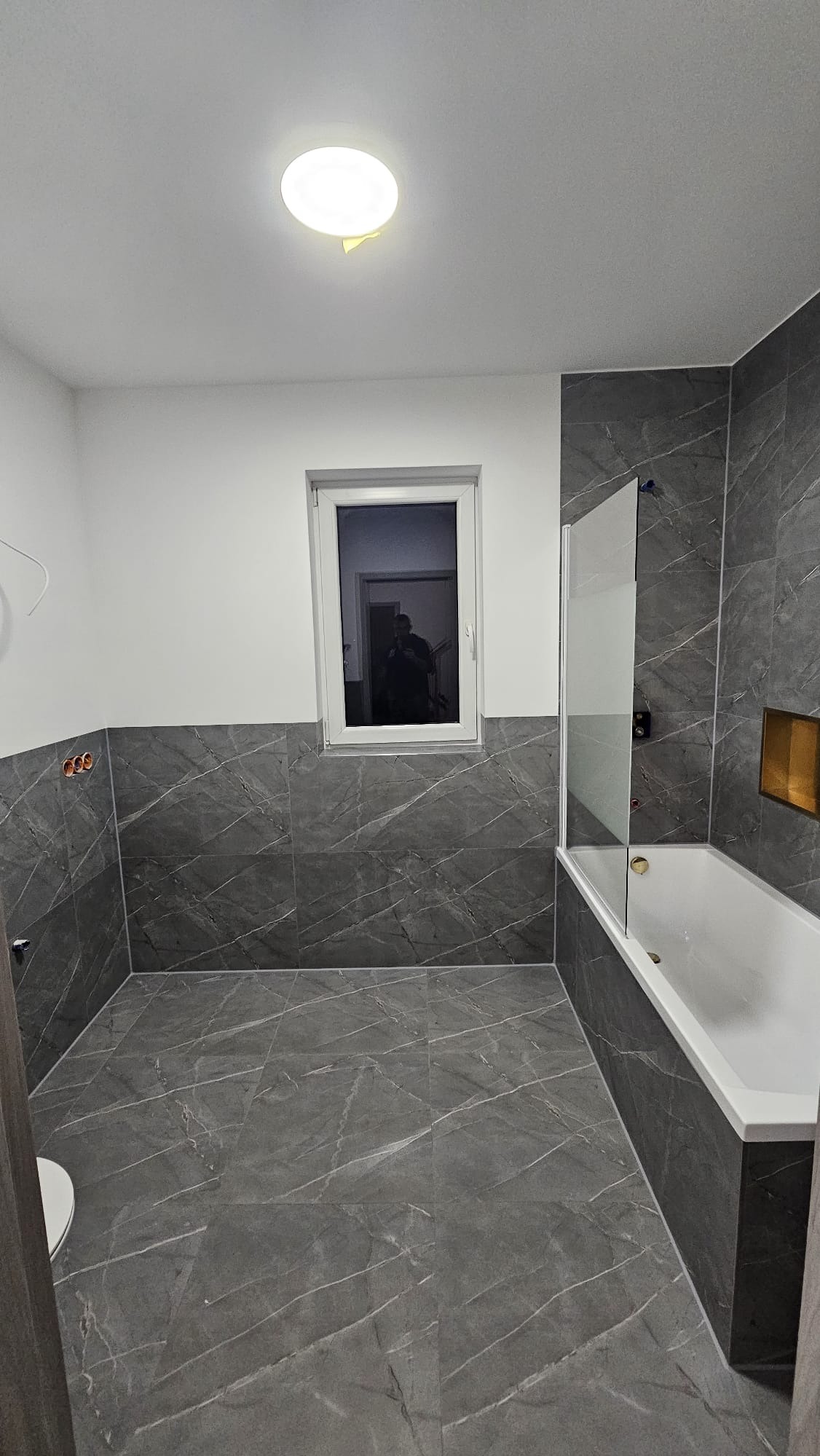 Wykończona łazienka z szarymi płytkami imitującymi marmur, białą wanną z parawanem i oknem z odbiciem osoby. Nowoczesne oświetlenie sufitowe.