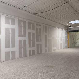 Piotr Dziuba - Wnętrze w trakcie prac wykończeniowych. Widoczne ściany z nałożoną warstwą wyrównującą, przygotowane do dalszych prac. Jasne, przestronne pomieszczenie z widocznym oświetleniem.