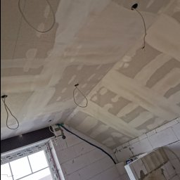 Piotr Dziuba - Skośny sufit po wstępnym wyrównaniu z widocznymi łączeniami, przygotowany do dalszych prac wykończeniowych. Widoczne kable elektryczne i okno z widokiem na budynek.