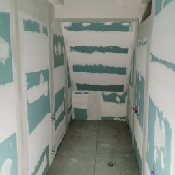 Piotr Dziuba - Wnętrze w trakcie prac wykończeniowych: ściany i sufit z widocznymi fragmentami zielonej płyty, częściowo pokryte białą warstwą. Na podłodze szare płyty.