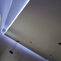 Piotr Dziuba - Nowoczesny sufit podwieszany z oświetleniem LED i punktowymi lampami, widziany pod kątem. Minimalistyczny design, gładka powierzchnia, subtelne światło.