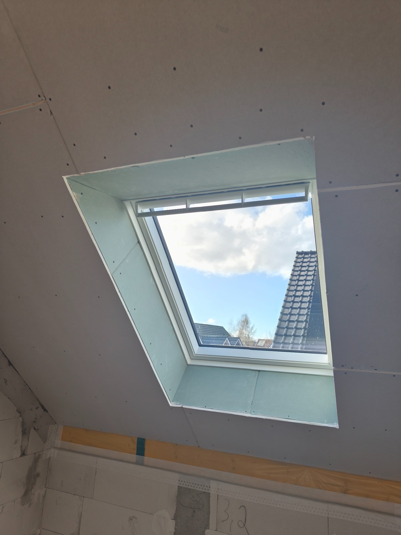 Montaż płyt na poddaszu wokół okna dachowego. Widoczne łączenia płyt i wkręty. W tle błękitne niebo i fragment dachu sąsiedniego budynku.