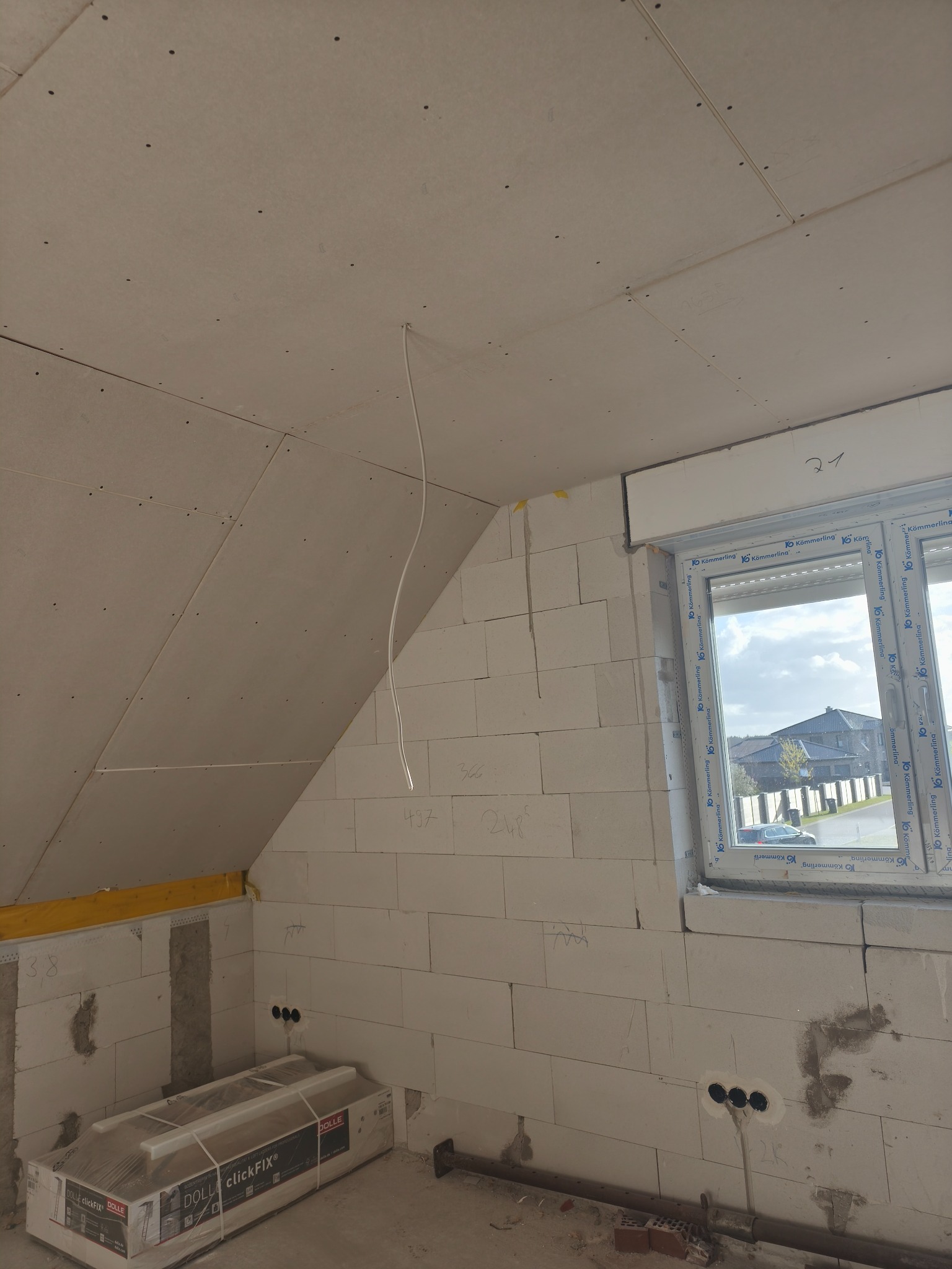 Montaż płyt w poddaszu: widoczne płyty na skosach i suficie, ściany z białego bloczka, okno z roletą. Na podłodze opakowanie płyt. Przewody elektryczne wystają ze ściany.