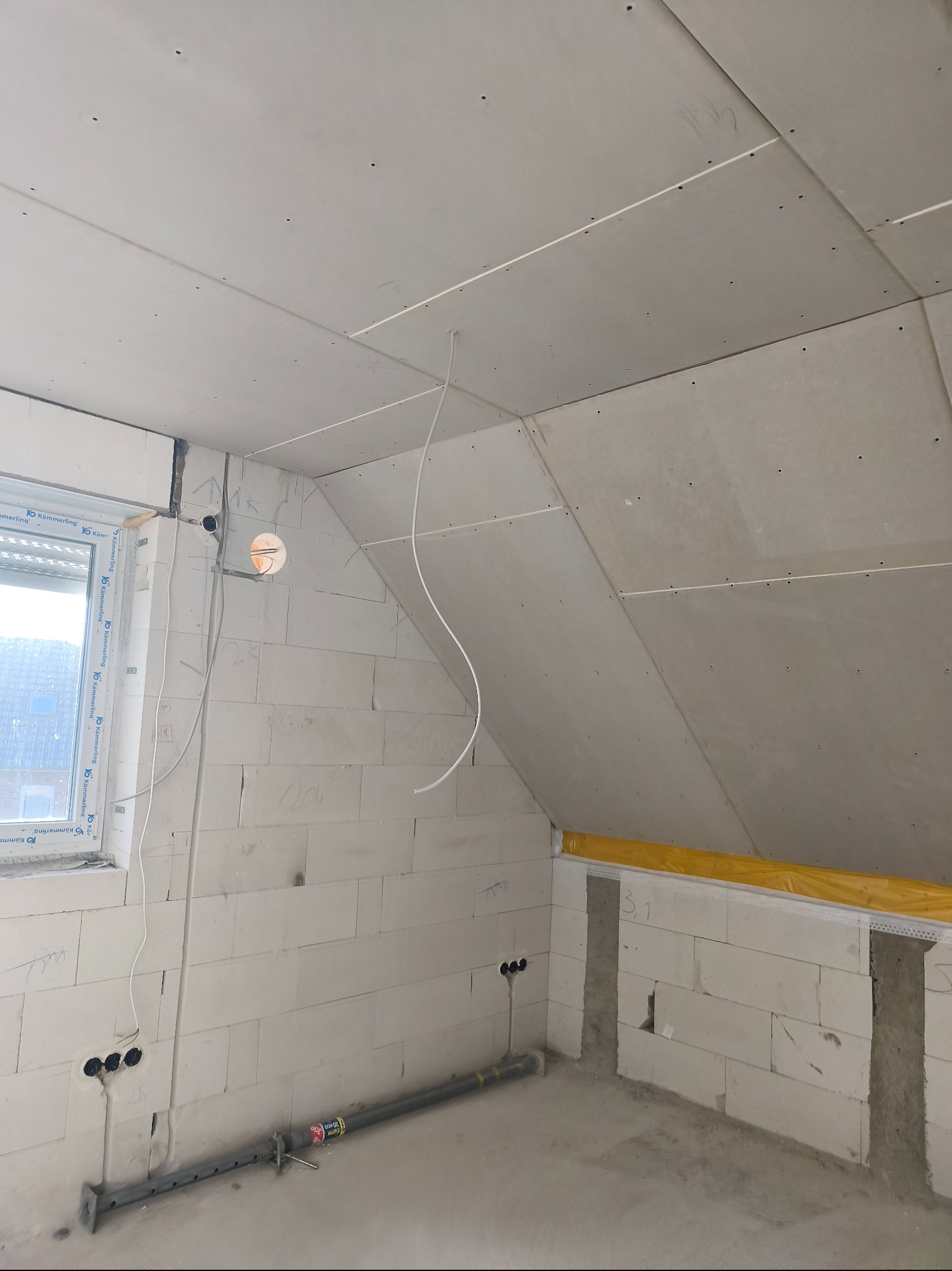 Poddasze w trakcie prac wykończeniowych: ściany z bloczków, zamontowane płyty na skosach, widoczne przewody elektryczne i okno. Etap przygotowania do dalszych prac.