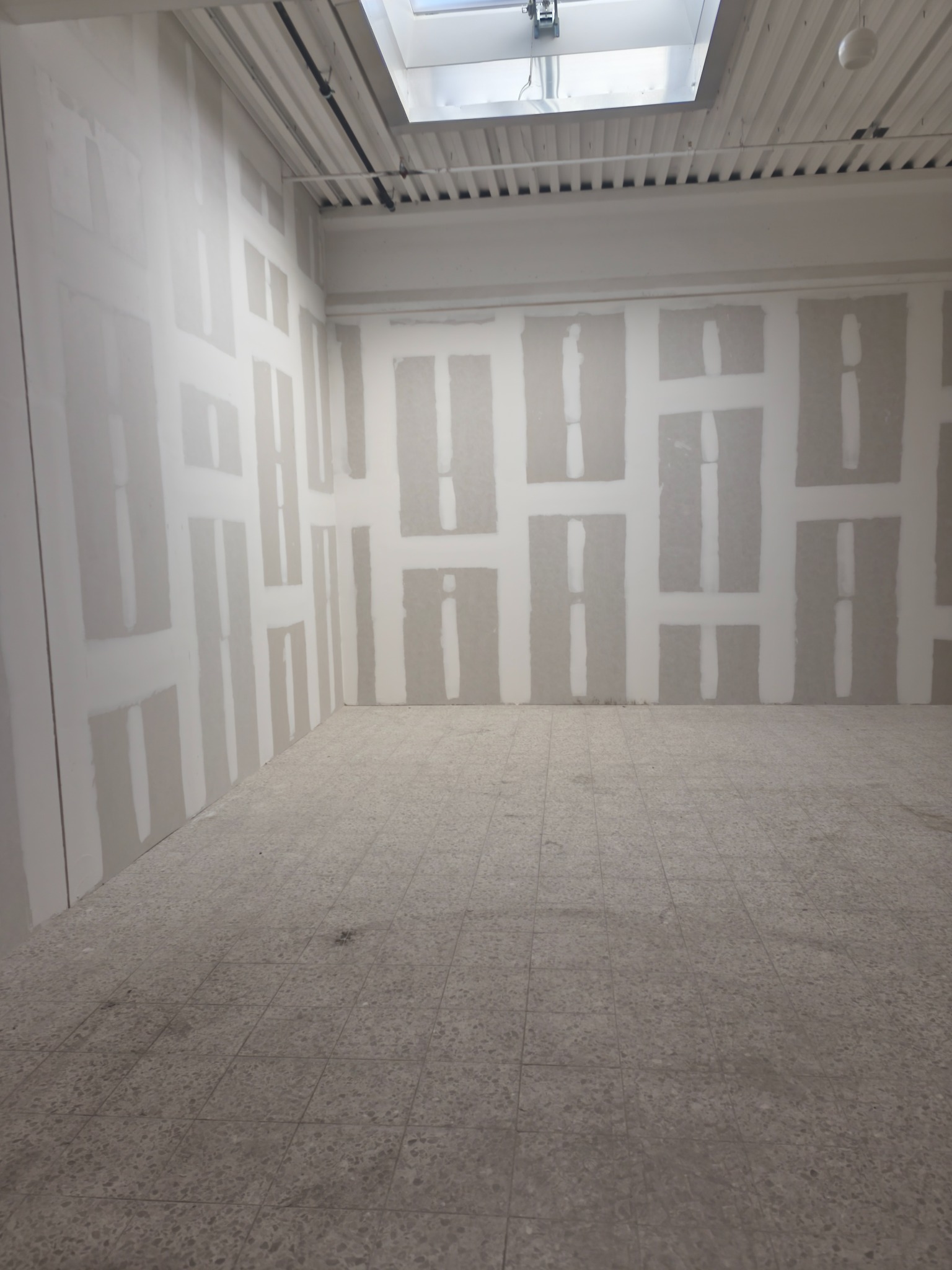 Wnętrze w trakcie prac wykończeniowych: ściany z fragmentami płyt, widoczne łączenia. Na suficie widoczny świetlik. Podłoga wyłożona płytkami.