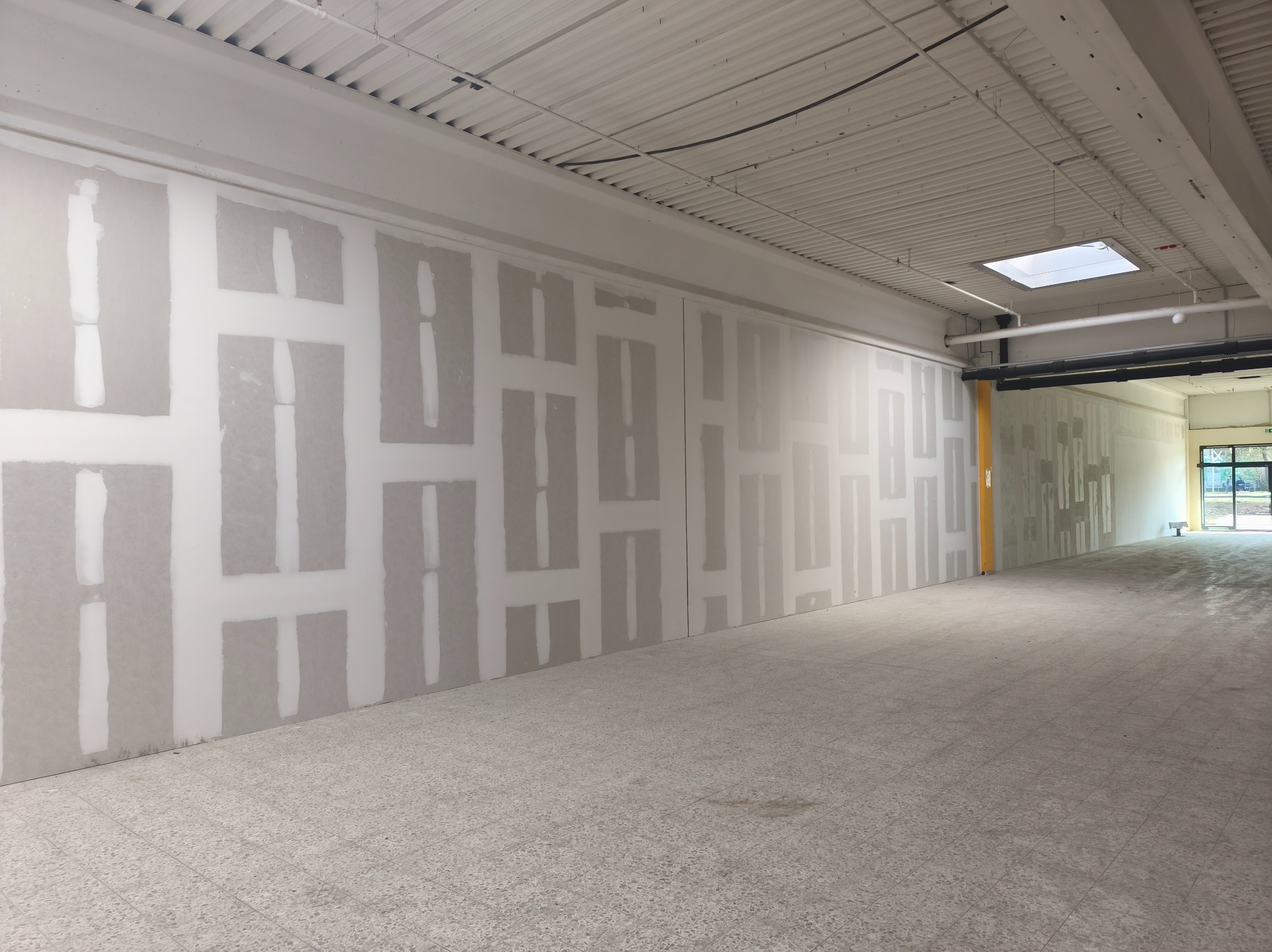 Wnętrze w trakcie prac wykończeniowych. Widoczne ściany z nałożoną warstwą wyrównującą, przygotowane do dalszych prac. Jasne, przestronne pomieszczenie z widocznym oświetleniem.