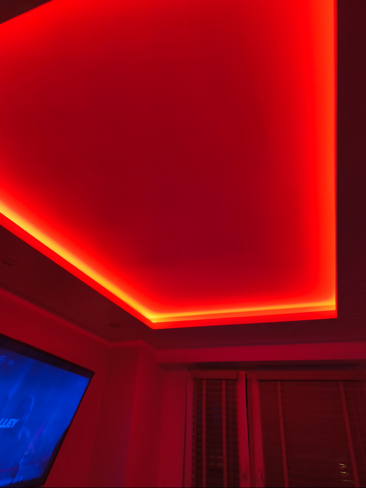 Podwieszany sufit z oświetleniem LED w odcieniach czerwieni i pomarańczy, tworzący nowoczesny efekt wizualny w pomieszczeniu. Widoczny fragment telewizora i okna.