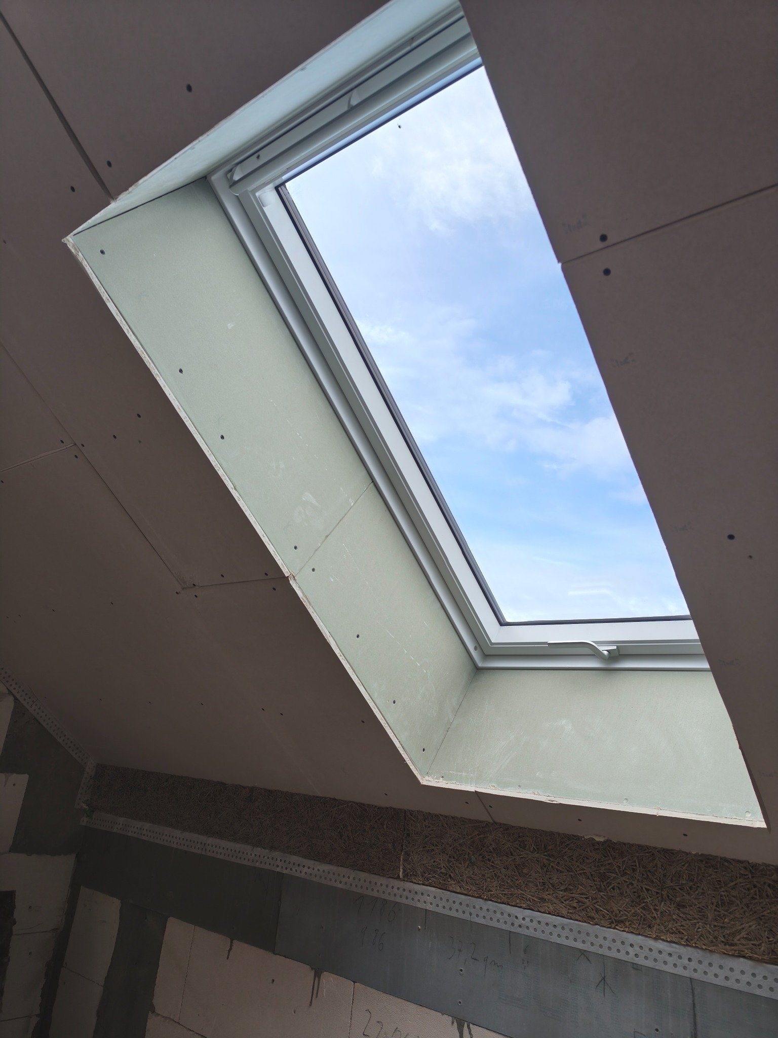 Montaż okna dachowego z obróbką płytami, widoczne łączenia i wkręty. W tle błękitne niebo przez okno. Ujęcie z dołu, podkreślające skos dachu.