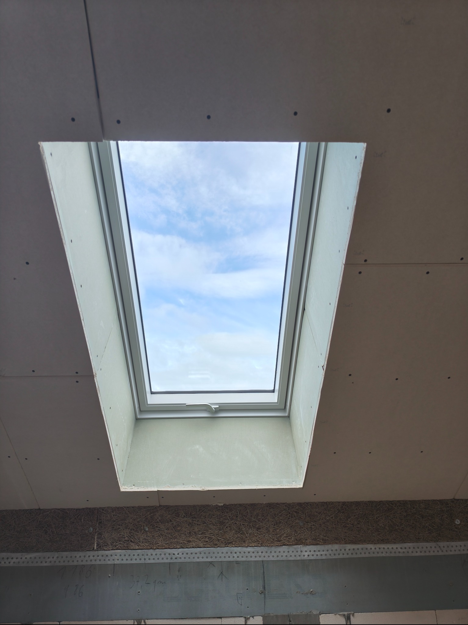 Montaż płyty karton gips wokół okna dachowego, widoczne łączenia płyt i wkręty. W tle błękitne niebo przez okno.