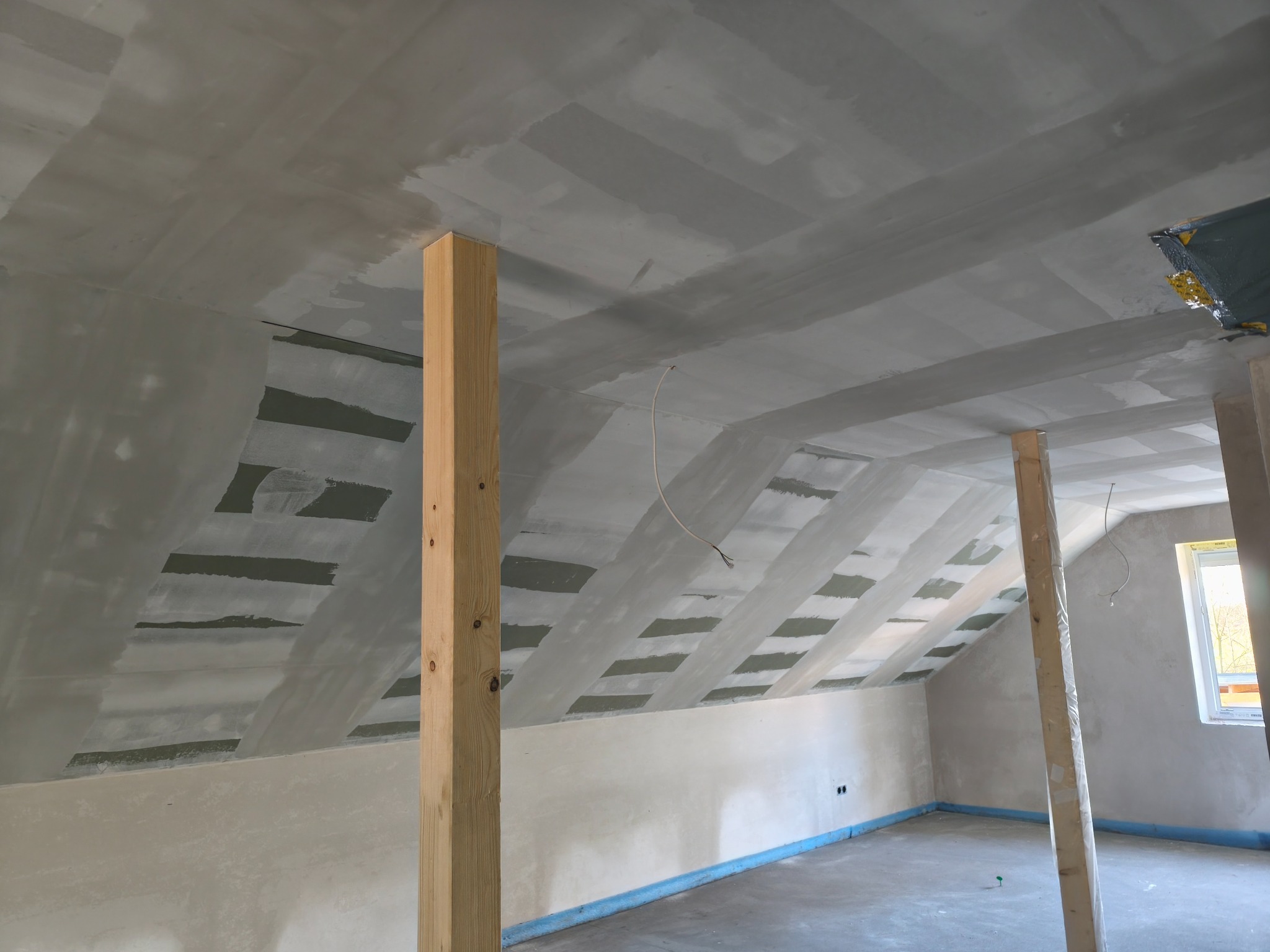 Sufit poddasza po wstępnym wyrównaniu w Jastrowie. Widoczne drewniane słupy i fragment okna. Ściany przygotowane do dalszych prac wykończeniowych.