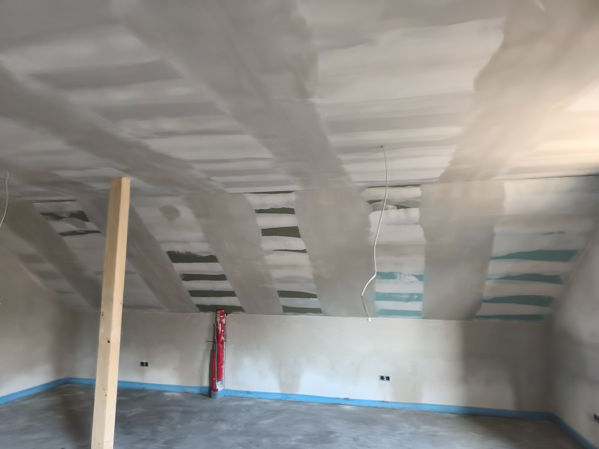 Poddasze w trakcie prac wykończeniowych: widoczne łączenia płyt na skosach sufitu, częściowo pokryte masą szpachlową, drewniany słup podtrzymujący konstrukcję.