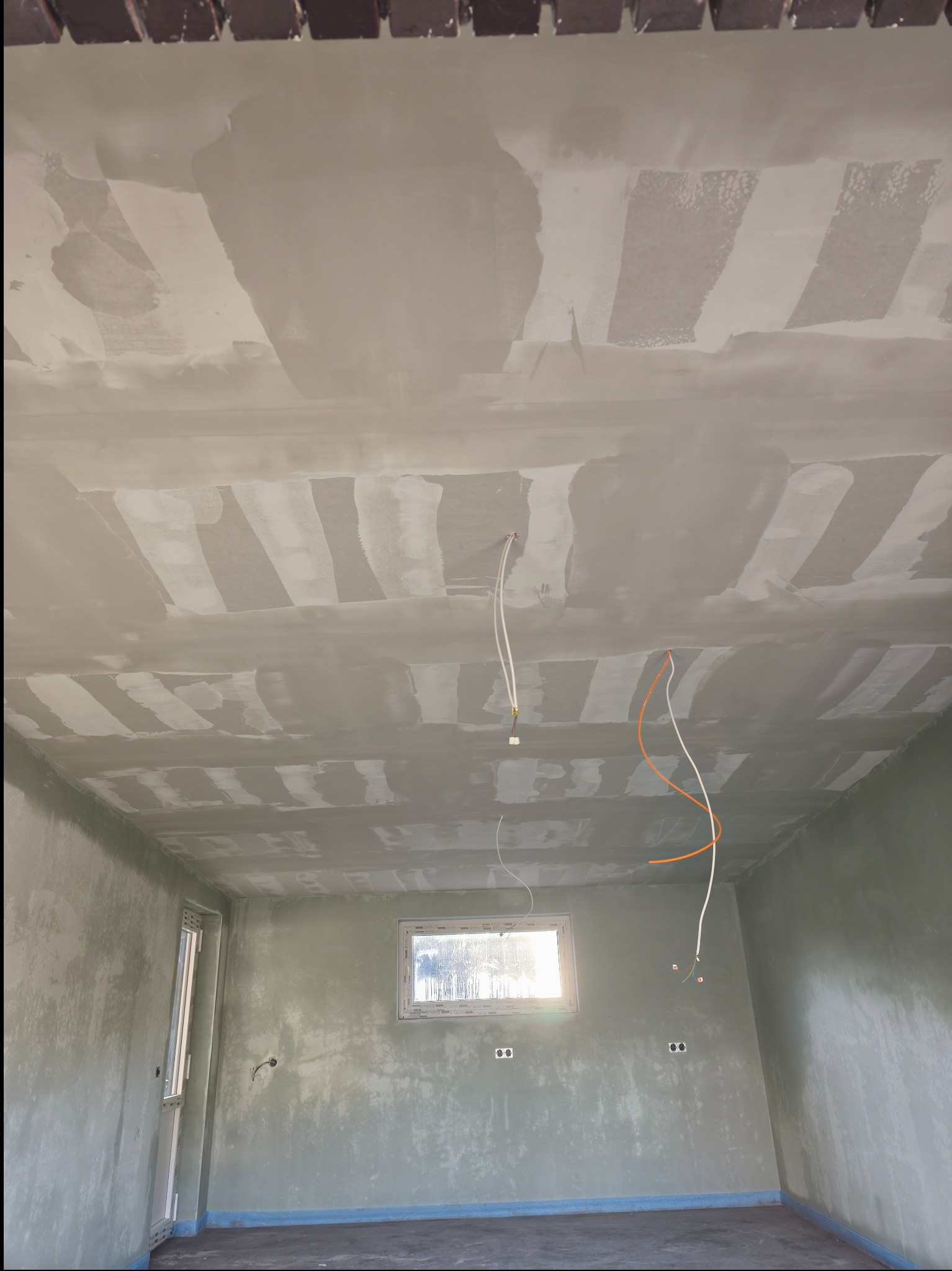 Wnętrze pokoju w trakcie prac wykończeniowych: surowy sufit z widocznymi łączeniami, ściany po wstępnym wyrównaniu, okno z widokiem na zewnątrz, wystające przewody.