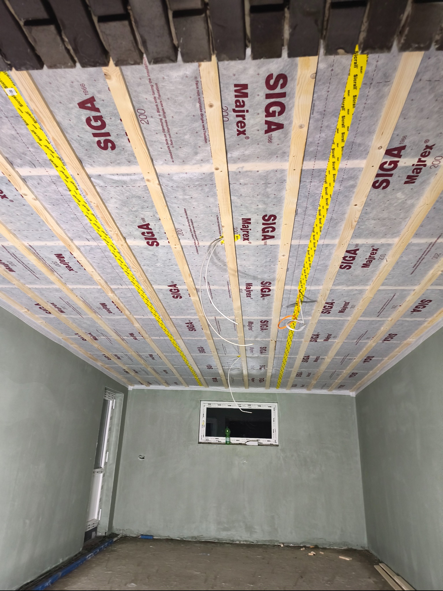 Montaż izolacji na drewnianej konstrukcji sufitu z widocznymi przewodami elektrycznymi i ścianami przygotowanymi do wykończenia. Folia izolacyjna z logo Siga.