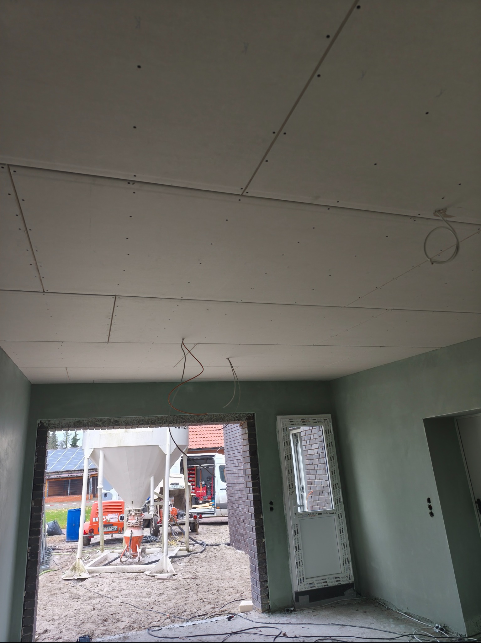 Wnętrze w trakcie prac wykończeniowych: zamontowane płyty na suficie, ściany przygotowane pod malowanie, widoczny sprzęt budowlany na zewnątrz. Surowy stan.