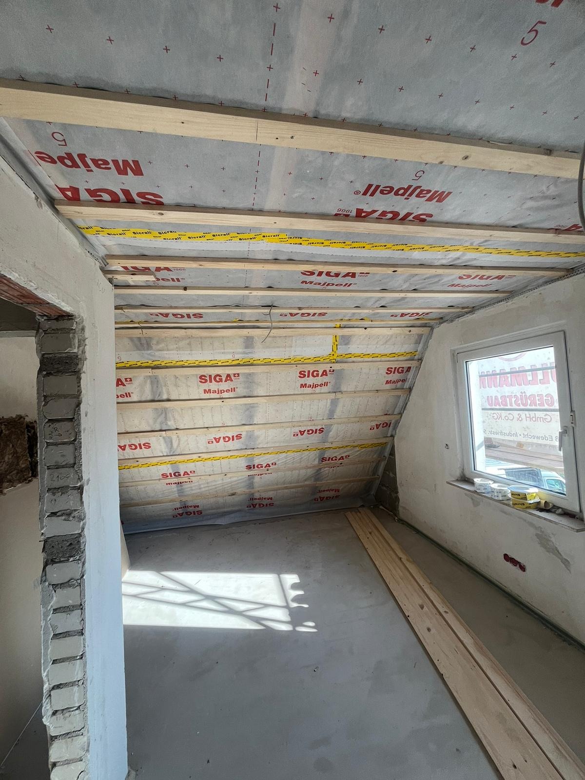 Poddasze w trakcie prac: drewniana konstrukcja, folia izolacyjna z nadrukiem, surowy beton na podłodze, okno. Widoczne materiały budowlane i elementy wykończeniowe.