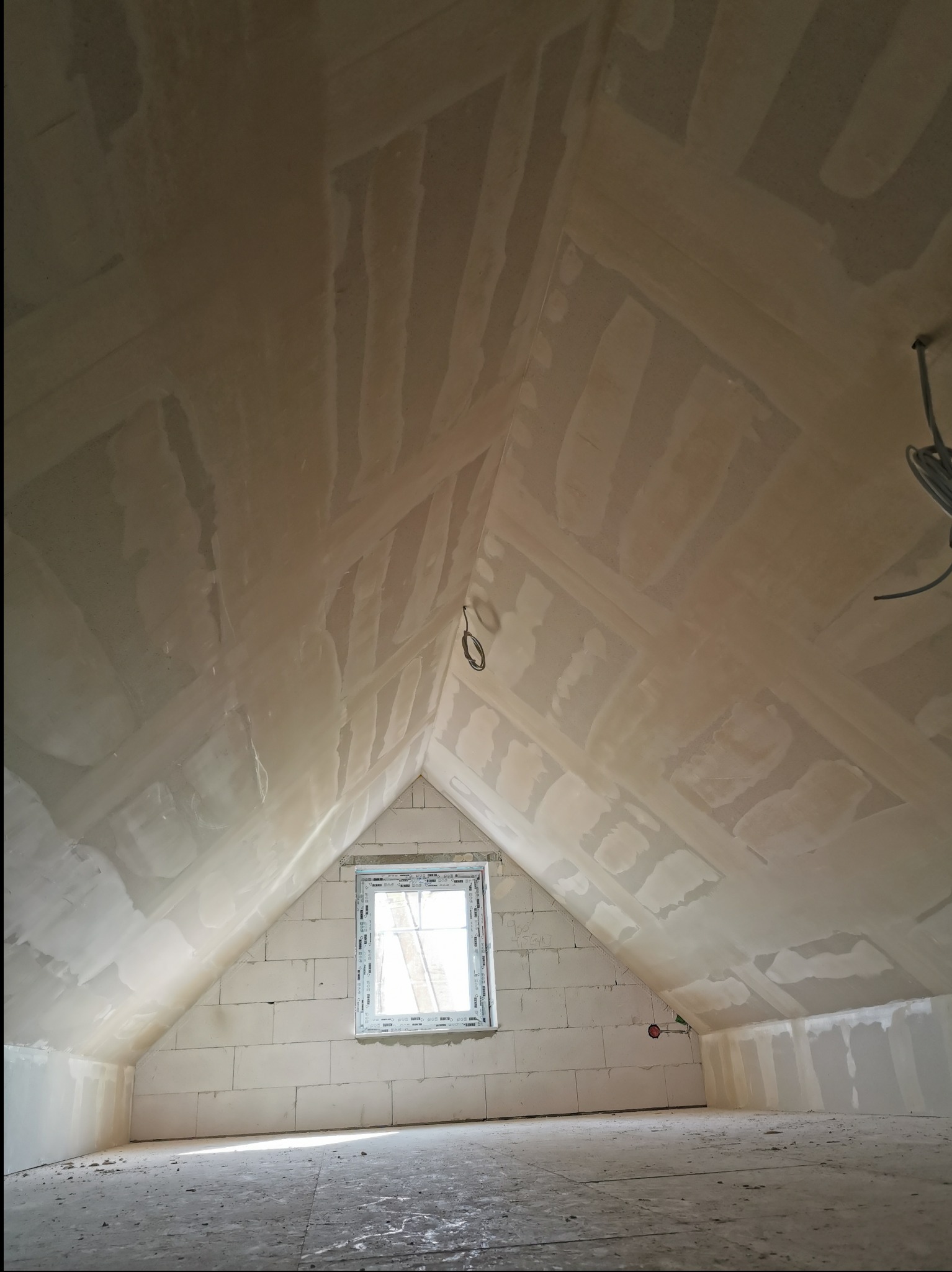 Poddasze po wstępnym wyrównaniu ścian i sufitu, widoczne łączenia płyt i niedoskonałości przed finalnym wykończeniem. Okno w szczycie doświetla wnętrze.