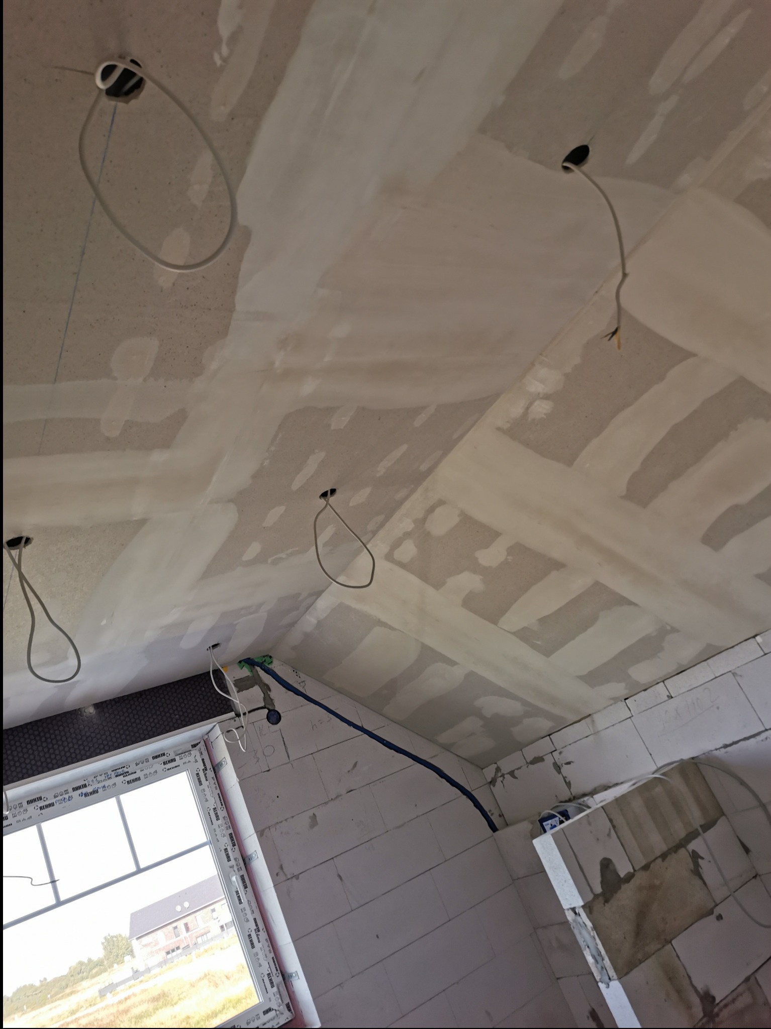 Skośny sufit po wstępnym wyrównaniu z widocznymi łączeniami, przygotowany do dalszych prac wykończeniowych. Widoczne kable elektryczne i okno z widokiem na budynek.