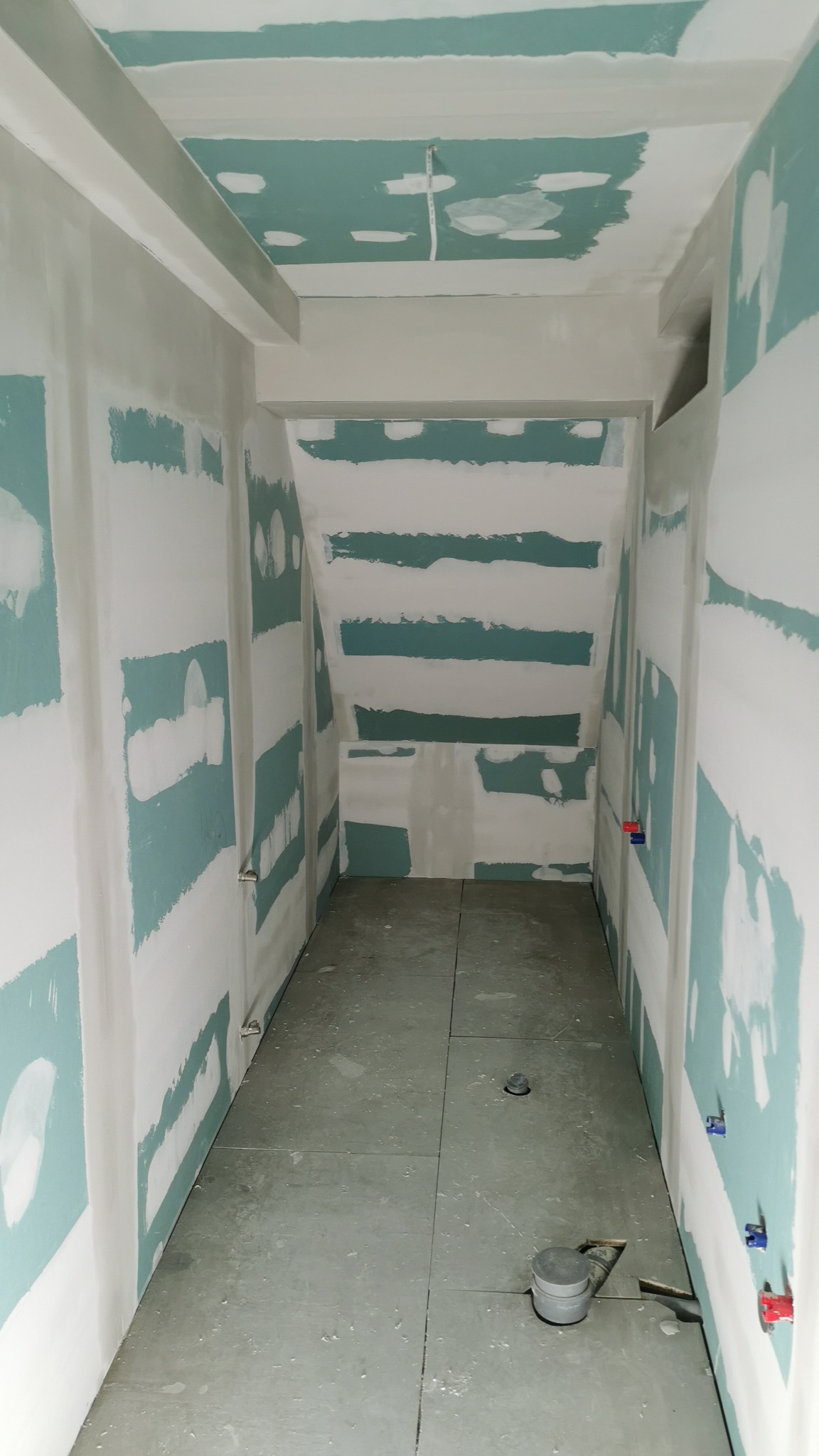 Wnętrze w trakcie prac wykończeniowych: ściany i sufit z widocznymi fragmentami zielonej płyty, częściowo pokryte białą warstwą. Na podłodze szare płyty.